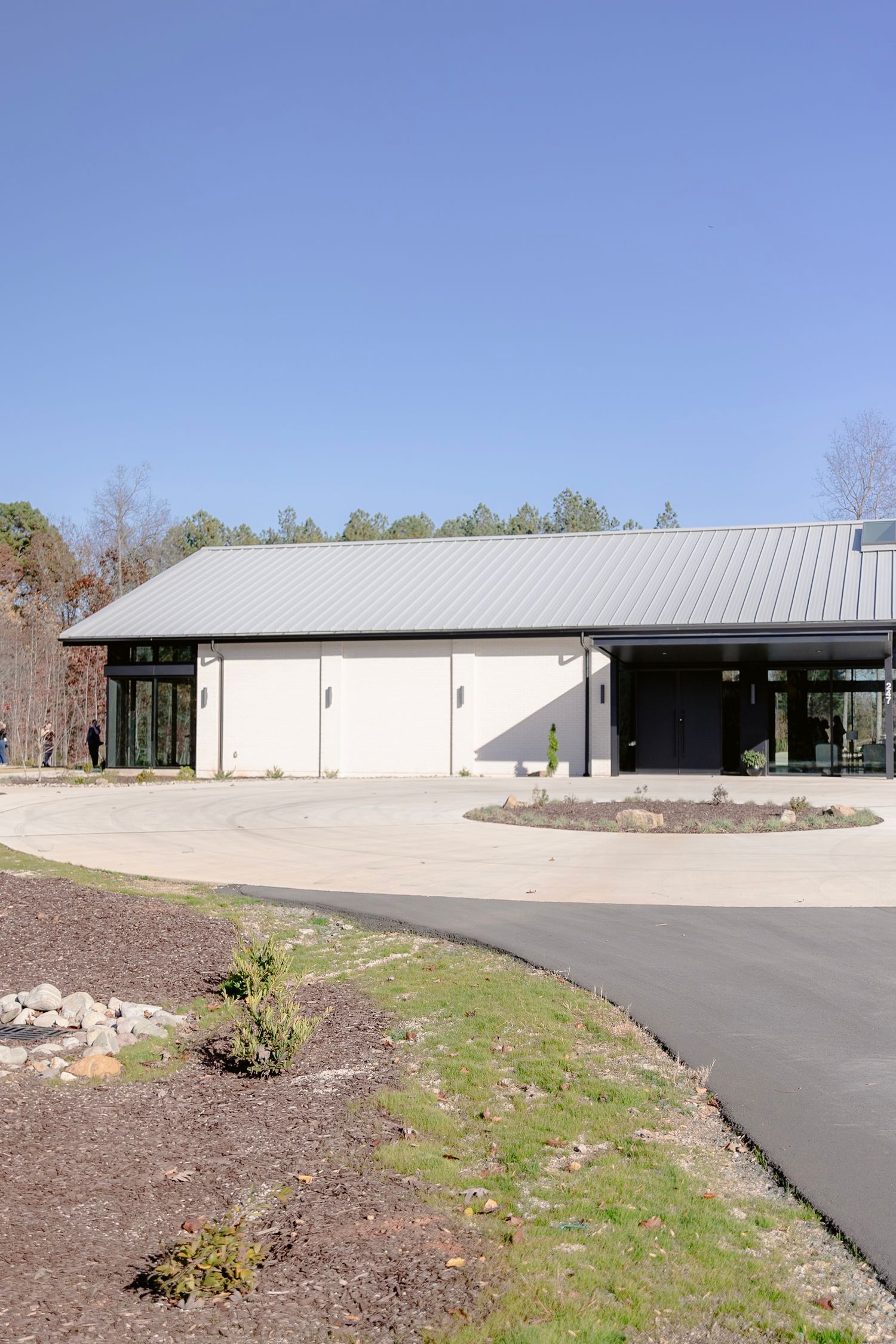Modern commercial building with white exterior, metal roof, and curved driveway surrounded by landscaped grounds.