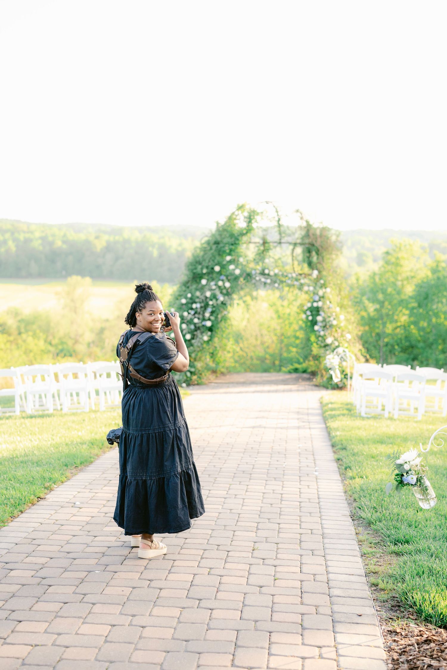 A toddler in a navy blue dress walks along a brick pathway surrounded by green grass and flowers on a sunny day.