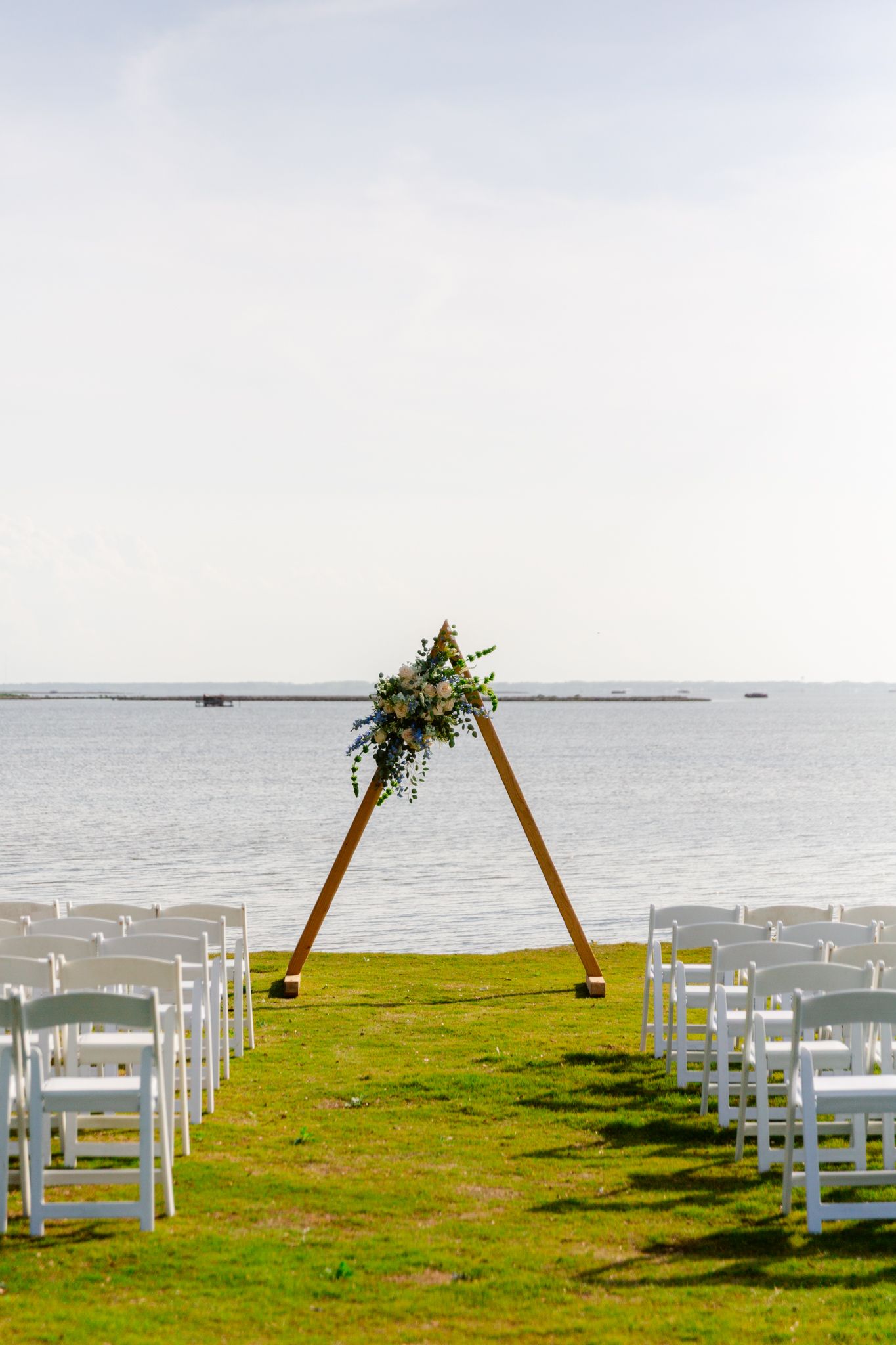 White chairs arranged for an outdoor wedding ceremony on a grassy cliff overlooking the ocean with a wooden arch.