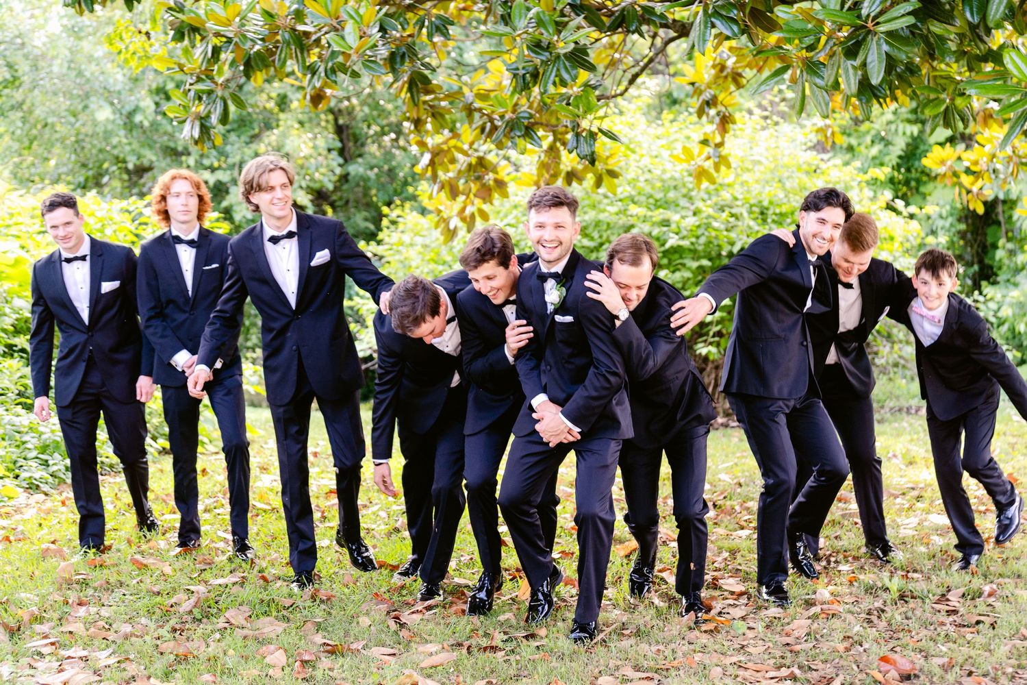 Wedding party in navy blue suits posing playfully and leaning to one side together outdoors under autumn trees.