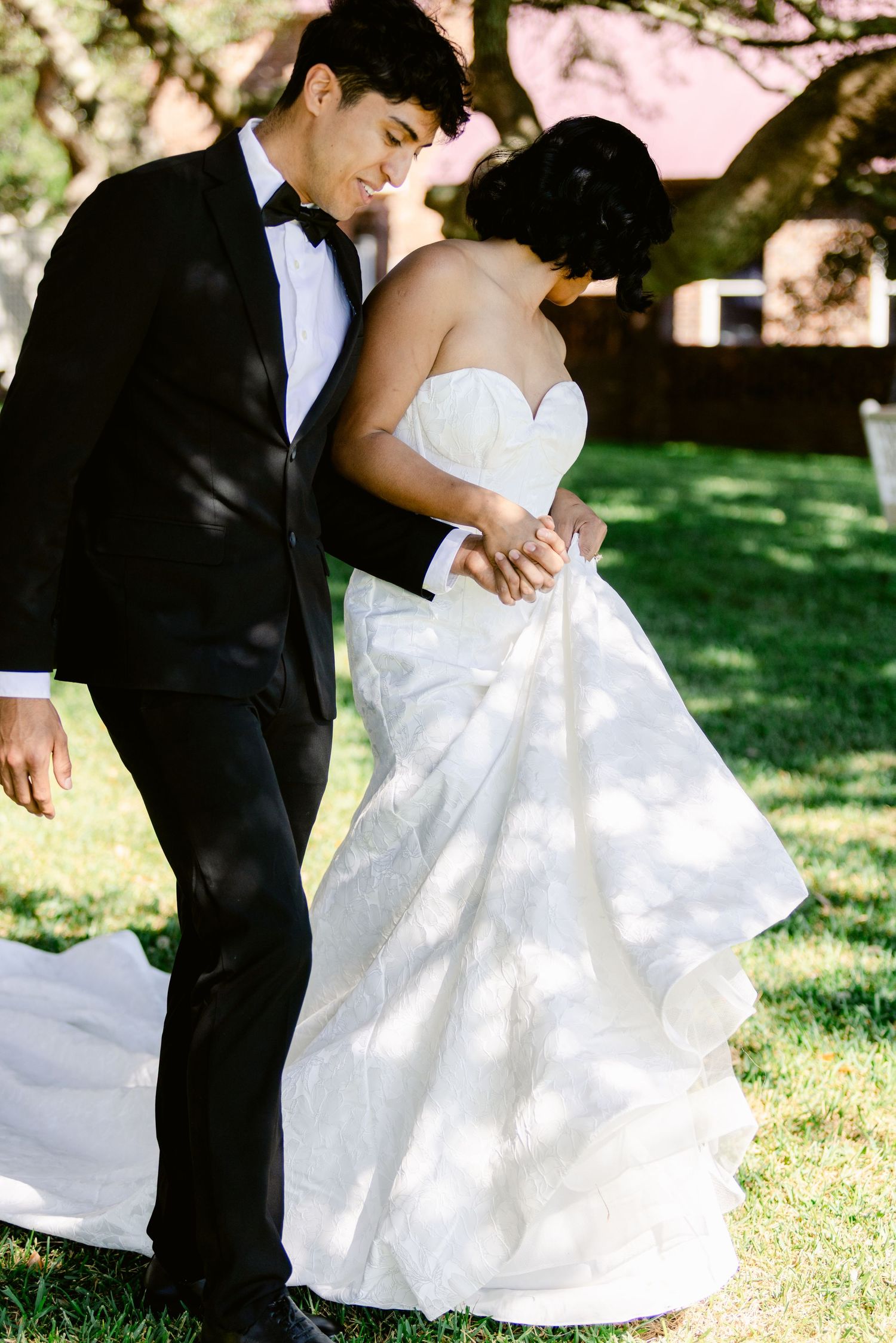 A couple in wedding attire dances together on a sunny lawn with dappled light filtering through trees.
