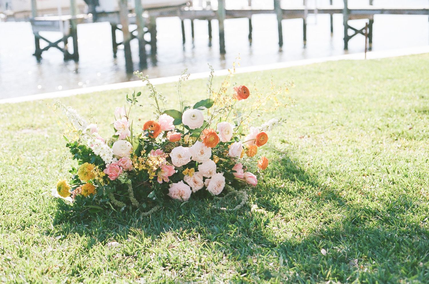 Lush floral arrangement with pink, peach and yellow blooms sits on grass near a waterfront dock.