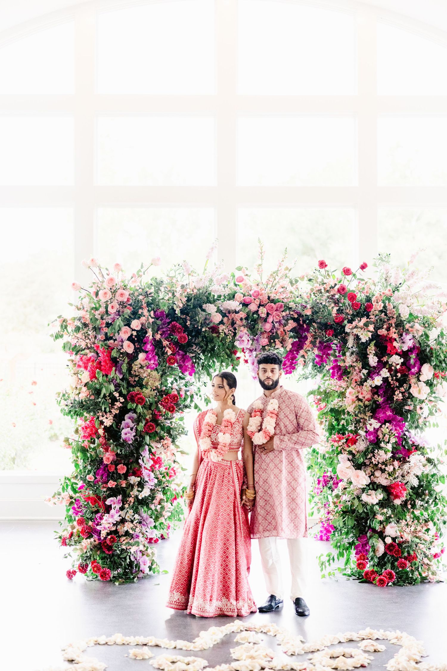 Couple in pink traditional Indian attire poses against a stunning floral backdrop with roses and greenery.