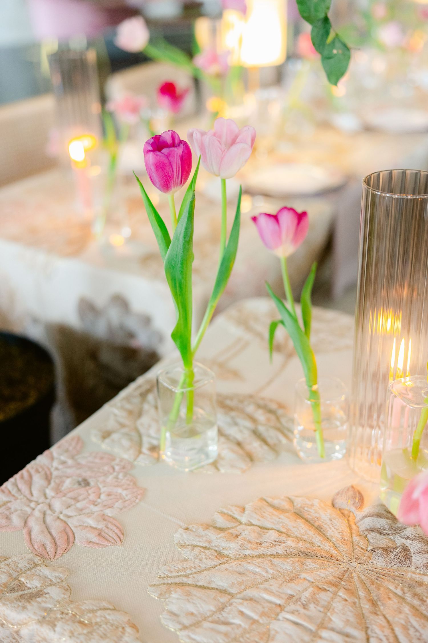 Pink tulips in glass vases on an elegant white textured tablecloth with glowing candlelight.