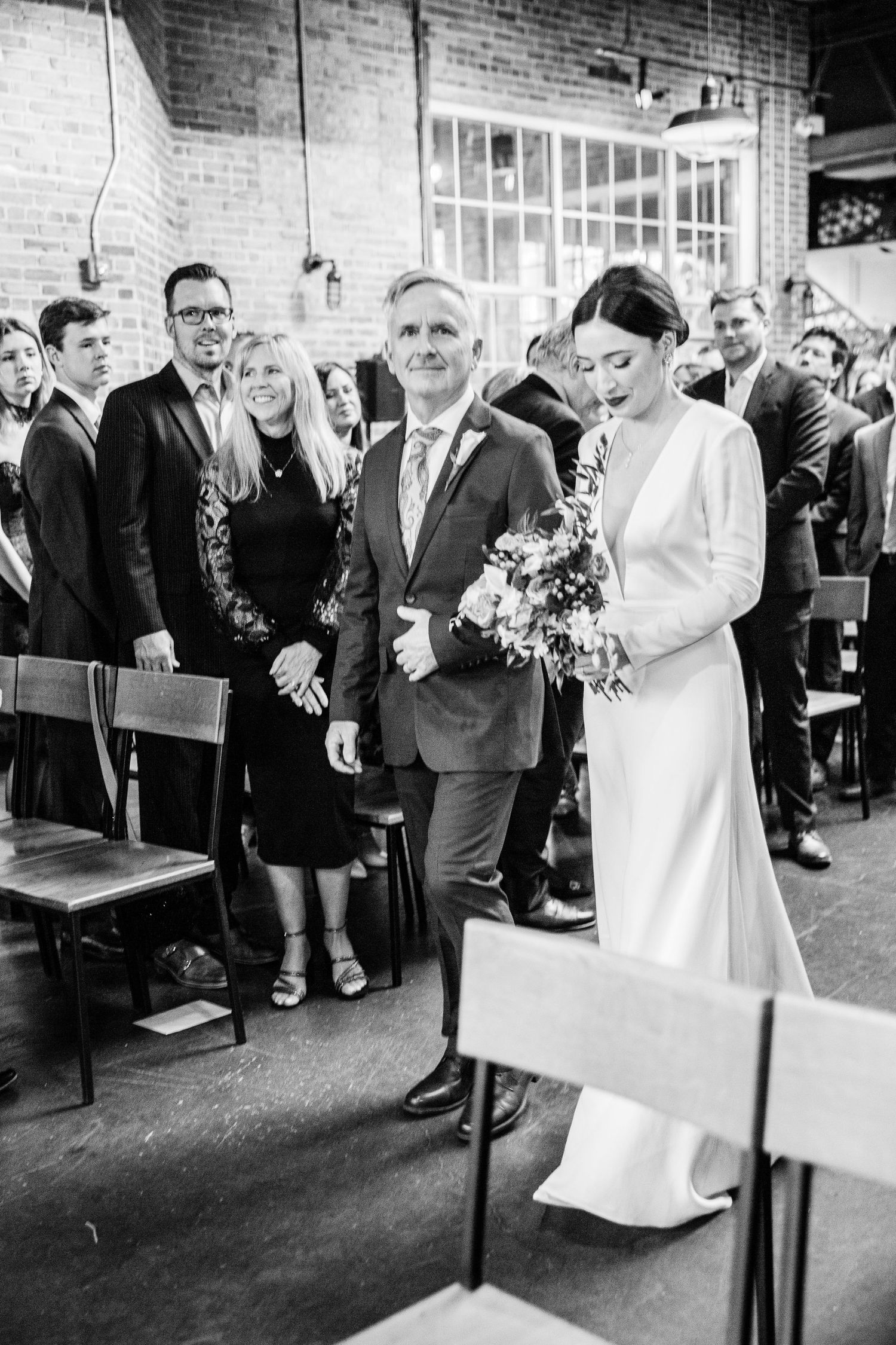 Black and white photo of a wedding ceremony in a brick-walled venue with guests watching from wooden chairs.