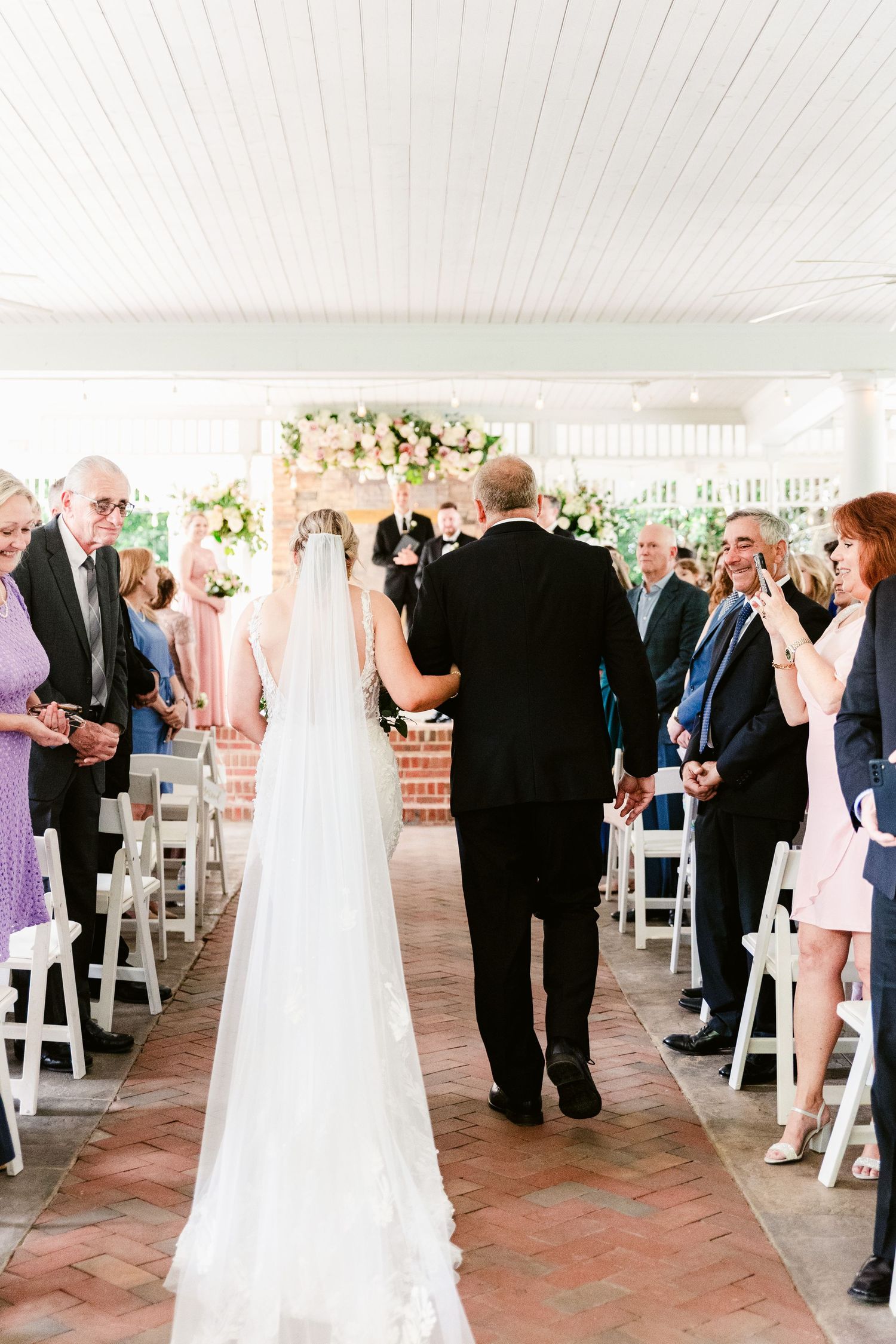 Wedding ceremony guests watch as a bride in white gown walks down a brick aisle under a covered porch.