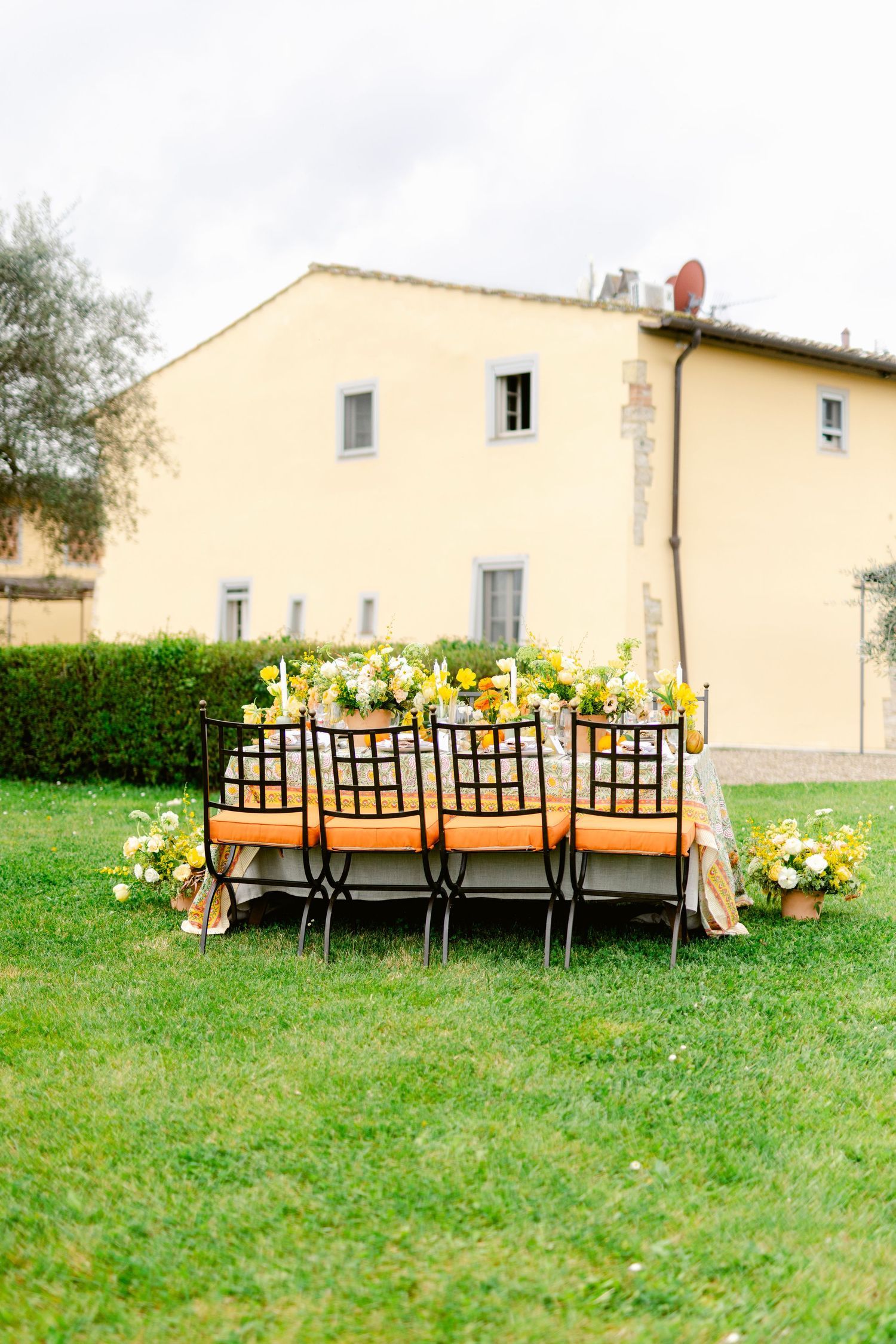 A long rustic wooden dining table with black chairs set up outdoors on a grassy lawn beside a yellow farmhouse.