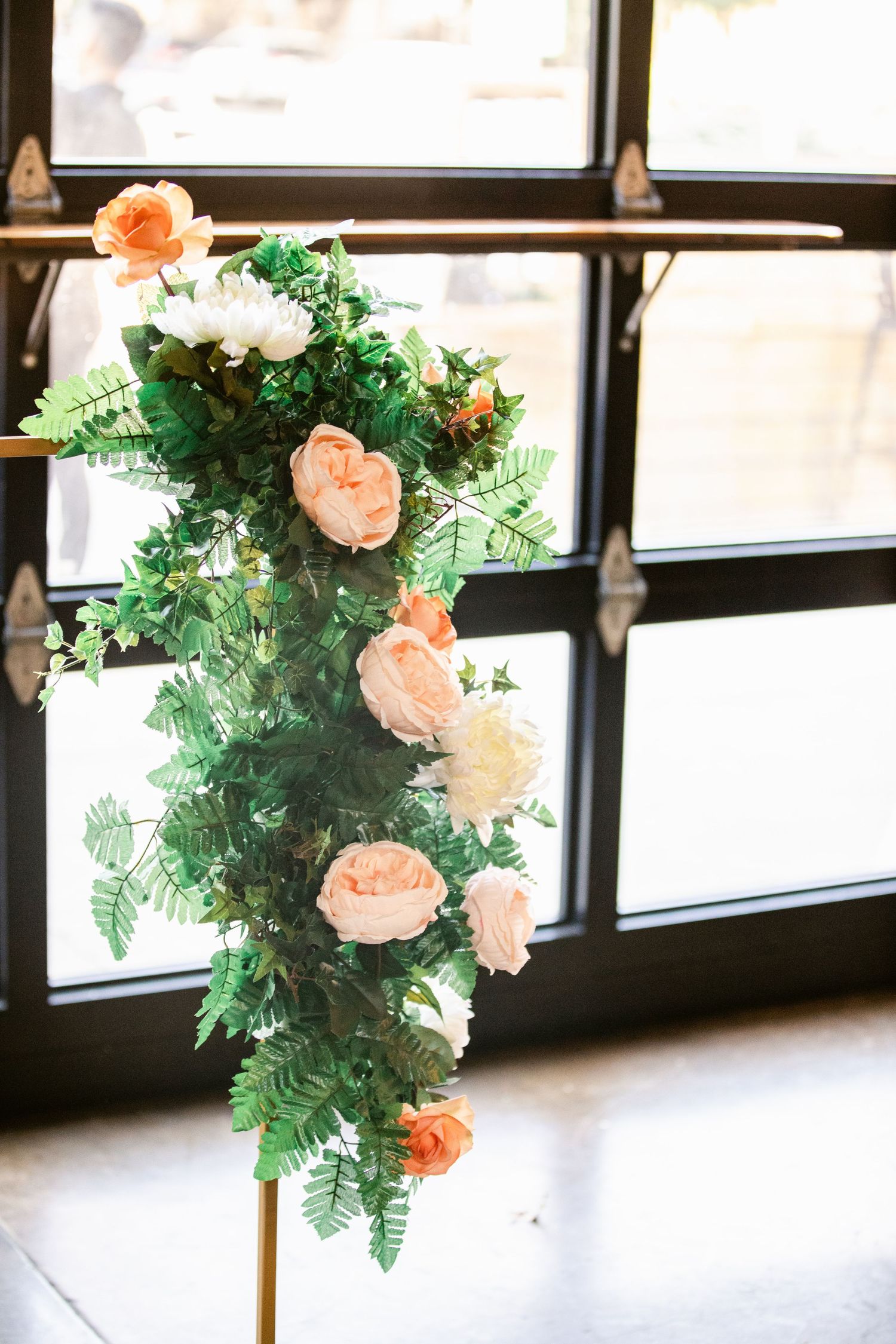 Elegant floral arrangement with peach roses and green ferns decorating an indoor venue with large windows.