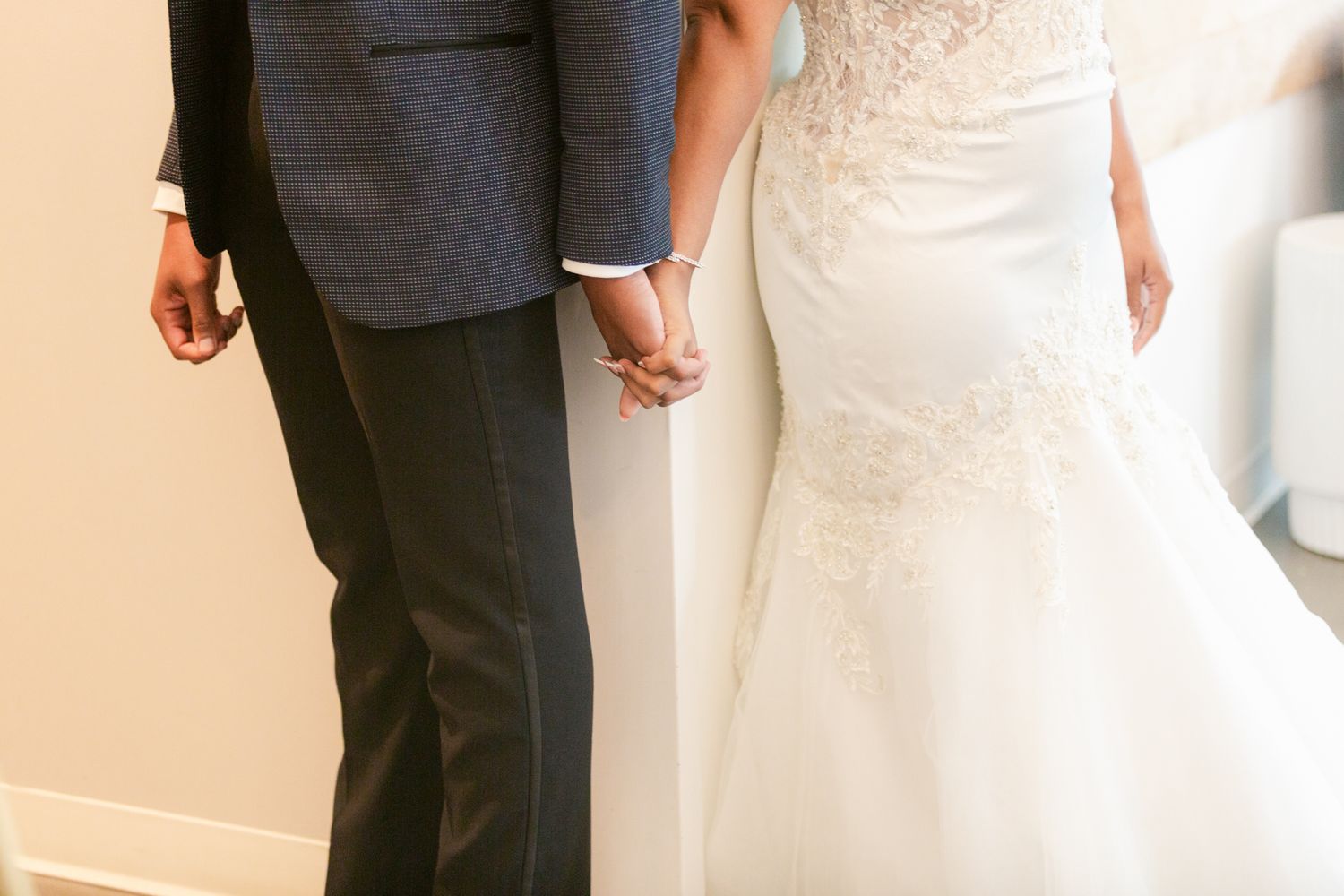 A close-up view of a bride and groom holding hands, showing her white lace dress and his navy suit.