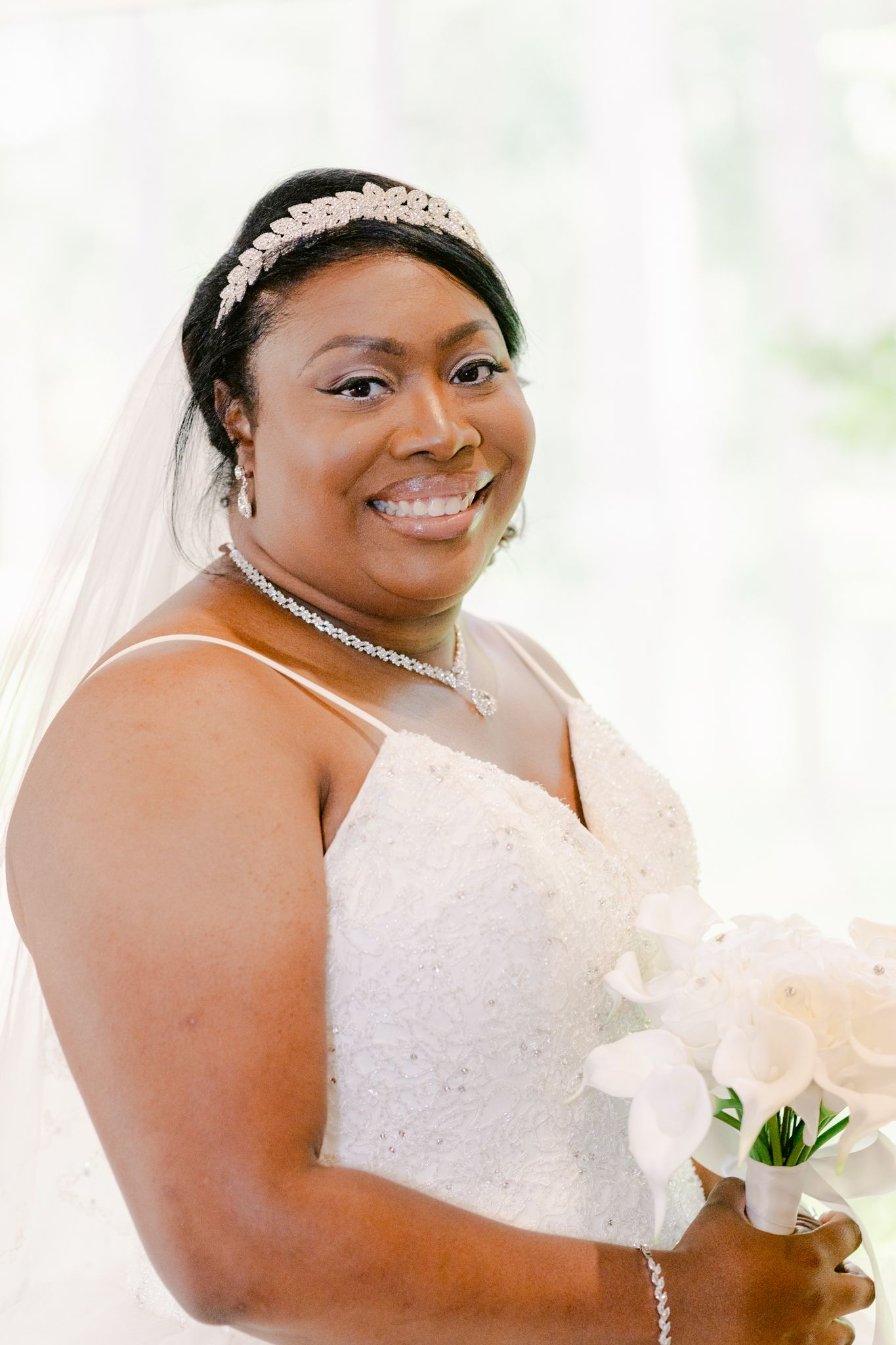 Beautiful bride in white lace dress and pearl tiara smiles radiantly in a bridal portrait series.