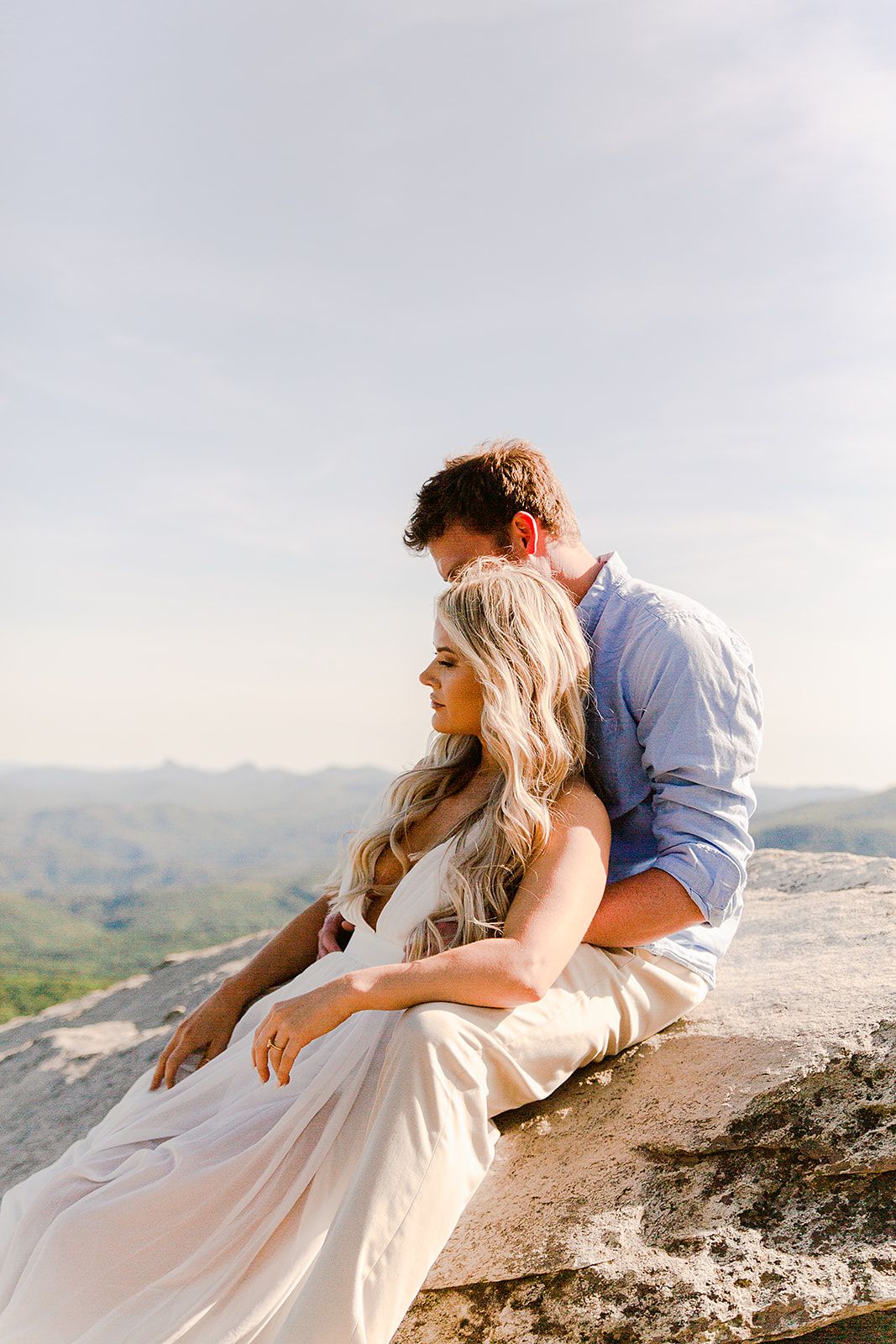 A romantic couple embraces on a mountain overlook with scenic rolling hills in the background during golden hour.