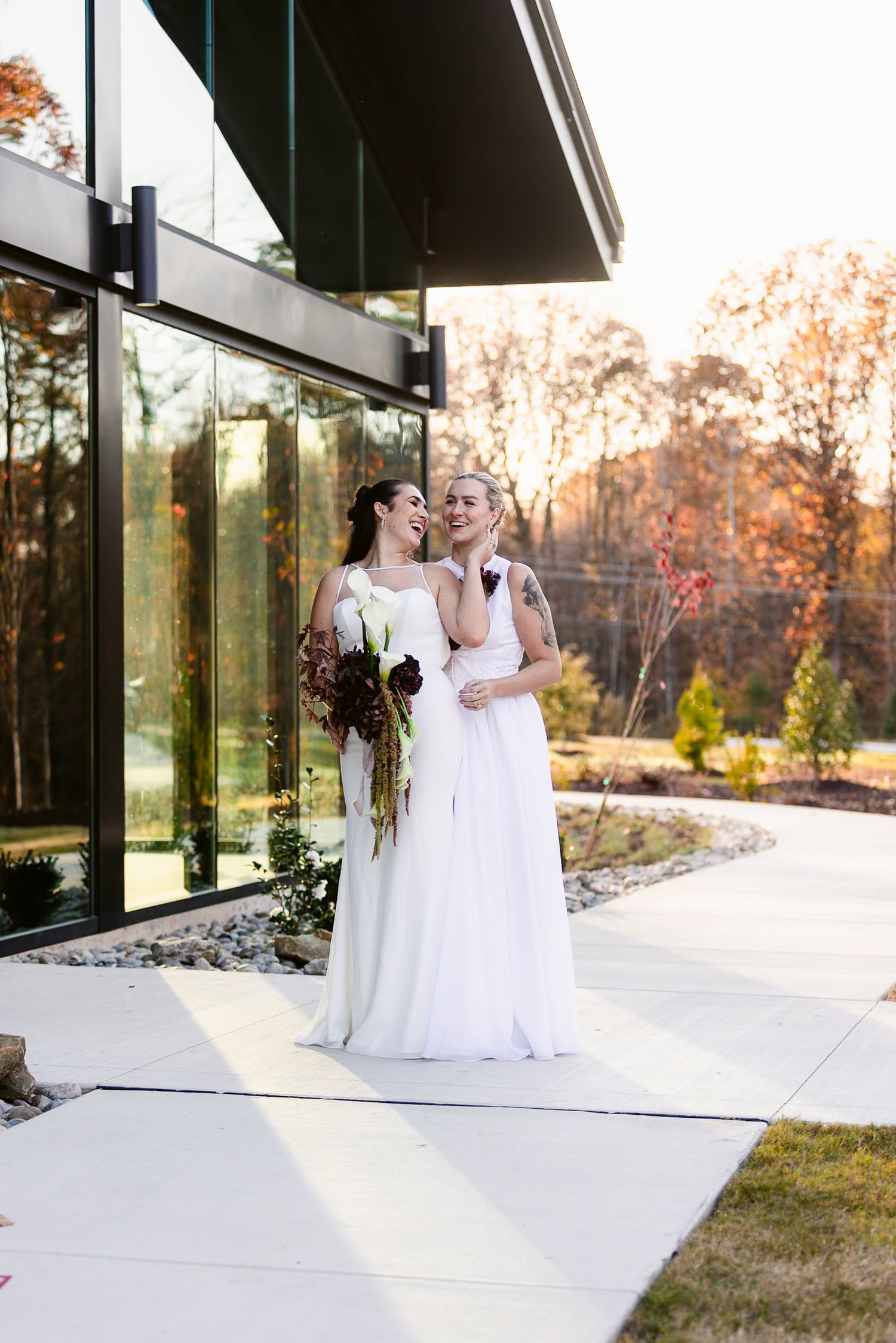 Two people in white wedding dresses share a tender moment on a modern patio with autumn foliage in the background.