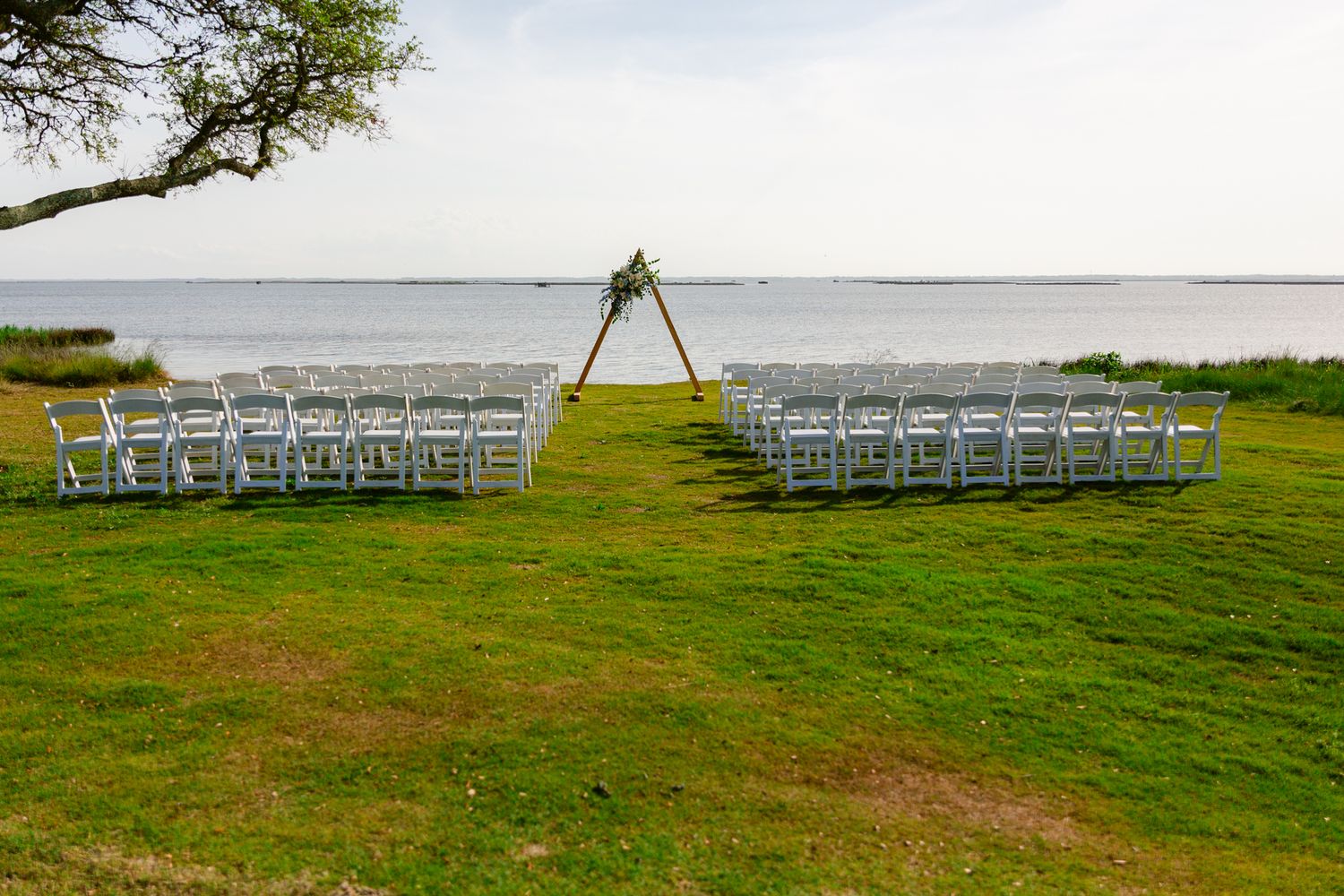 White wedding chairs arranged in rows on a green lawn overlooking a serene waterfront for an outdoor ceremony.
