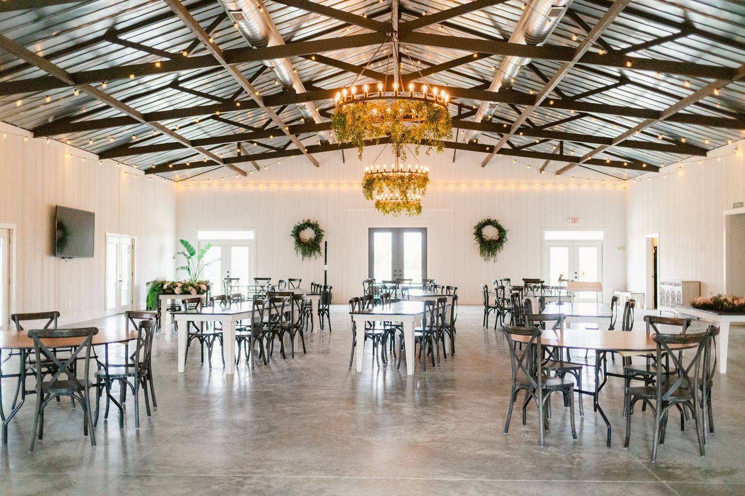 Large white reception hall with exposed wooden beams, chandeliers, and scattered tables and chairs for events.