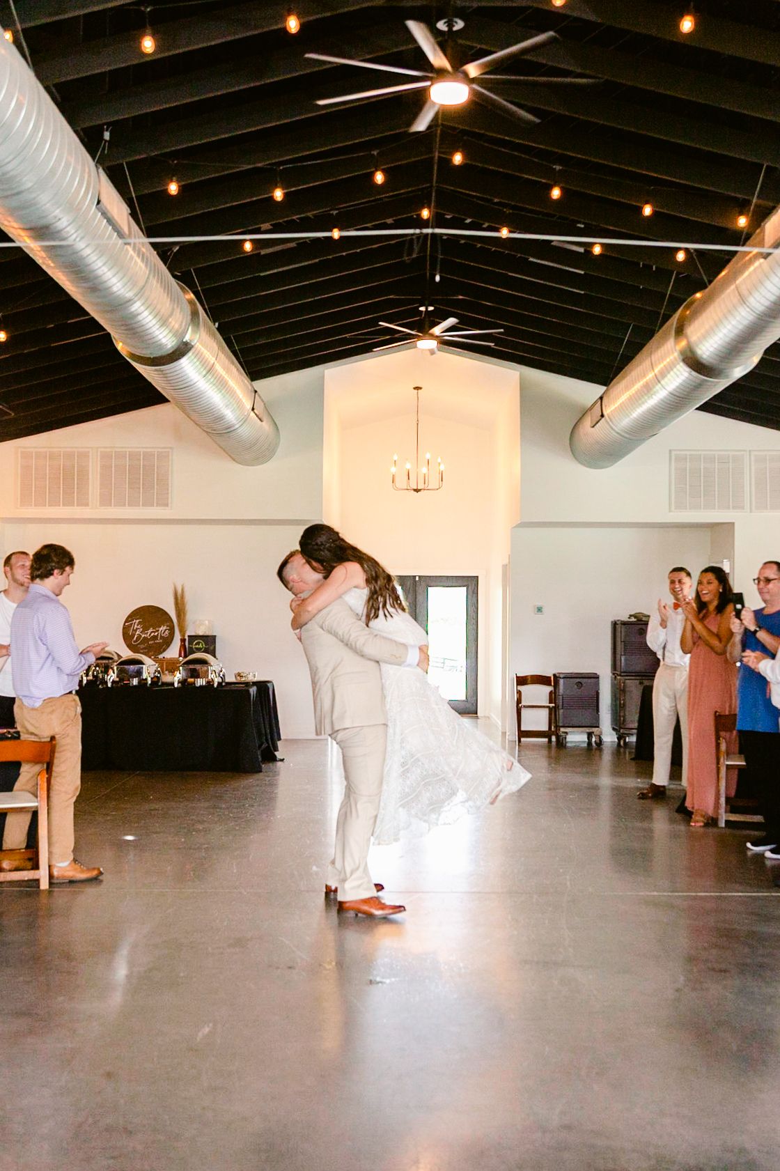 A spacious event venue with exposed ceiling beams, string lights, and people gathered in a modern industrial space.