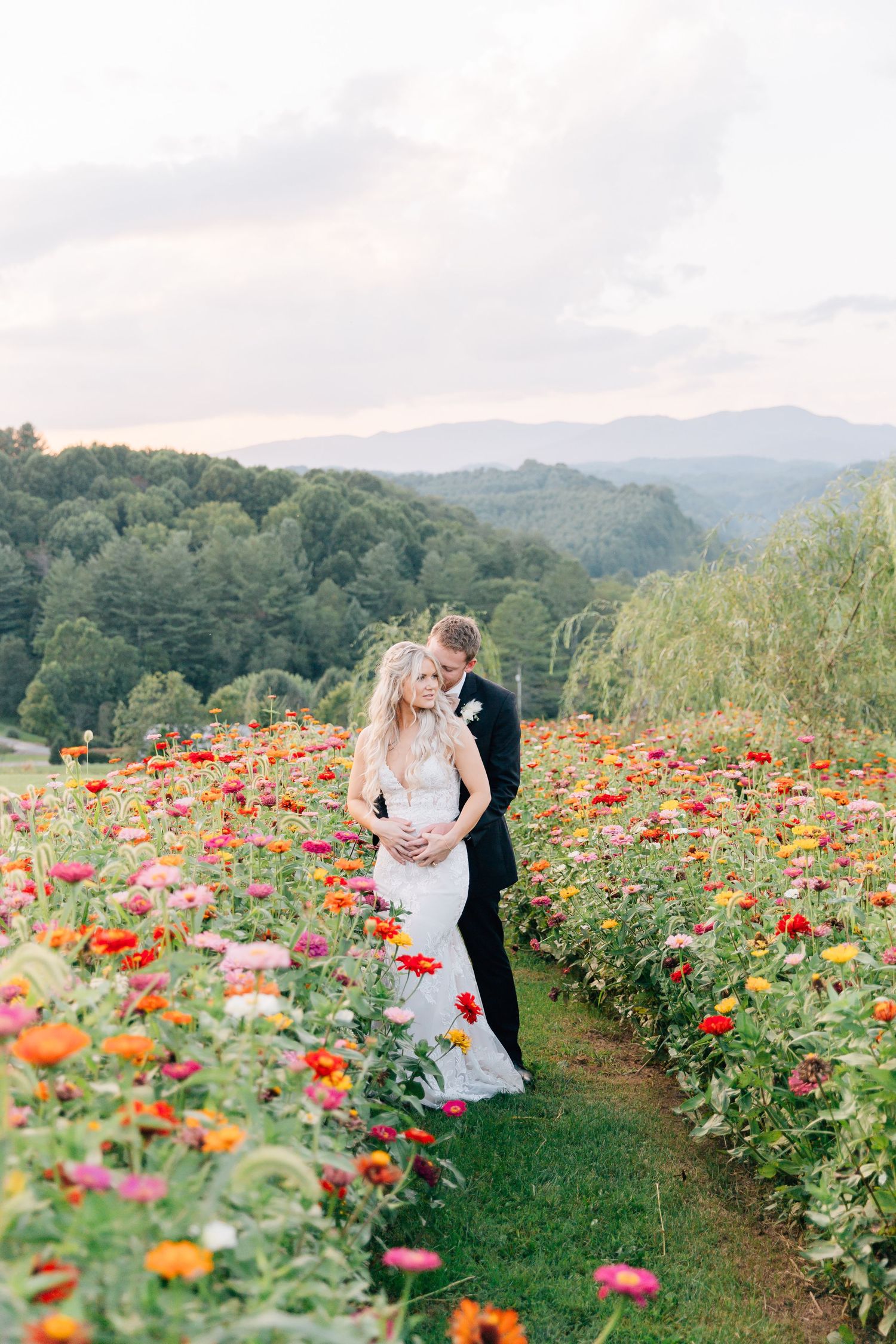 A couple embraces in a vibrant field of colorful zinnias with mountain views in the background.