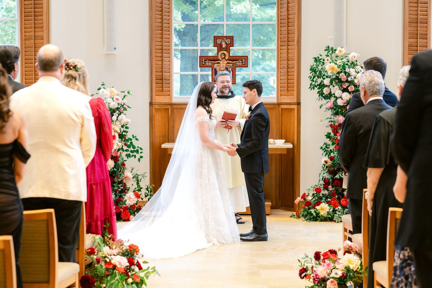 A wedding ceremony in a church with floral arrangements and a wooden cross at the altar.