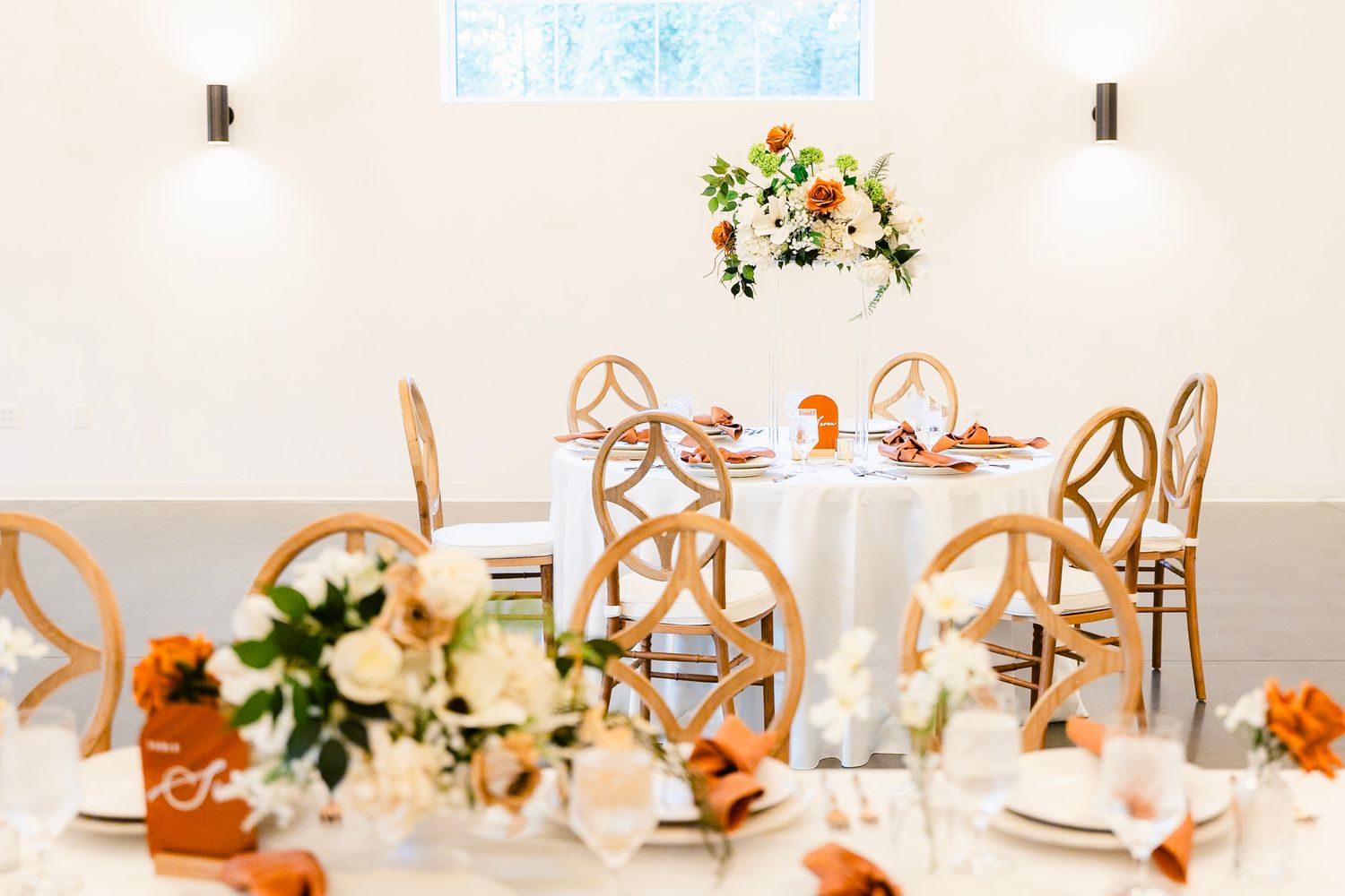 Elegant wedding reception table setting with wooden chairs and peachy floral centerpiece in a bright white room.