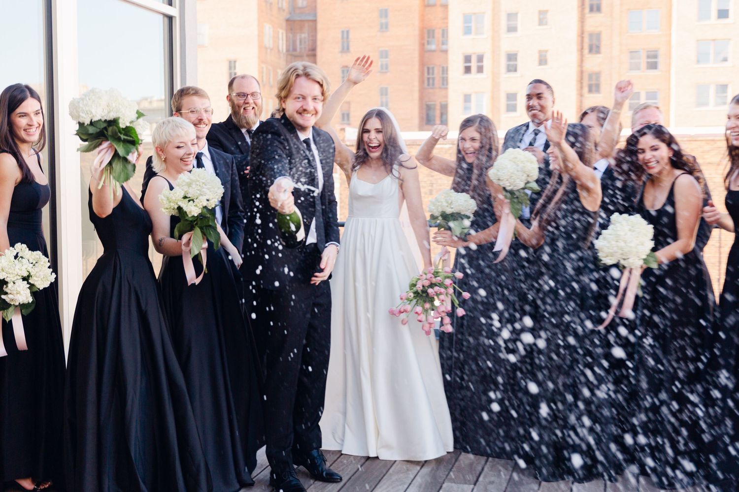 Wedding party celebrating with confetti on a rooftop, bridesmaids in black dresses.