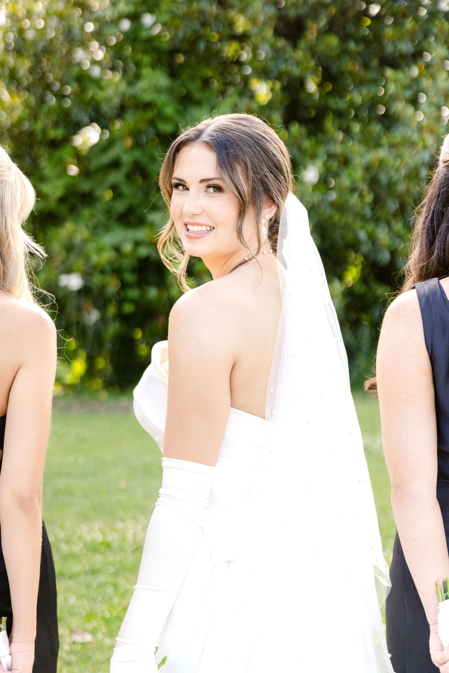 A bride in a white strapless wedding gown and veil poses outdoors with greenery in the background.