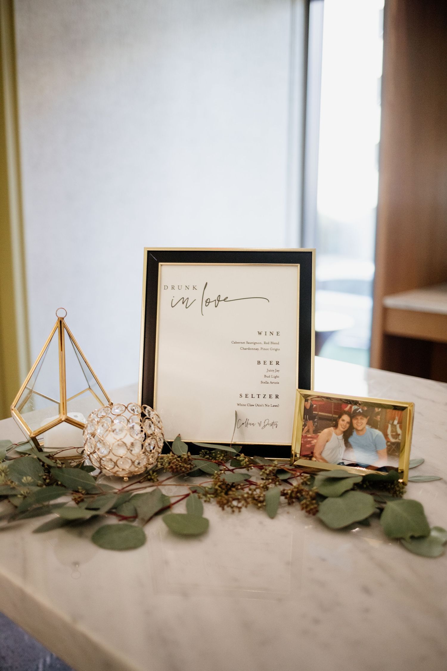 Wedding detail display with framed sign, geometric terrarium, and eucalyptus leaves on gray surface.