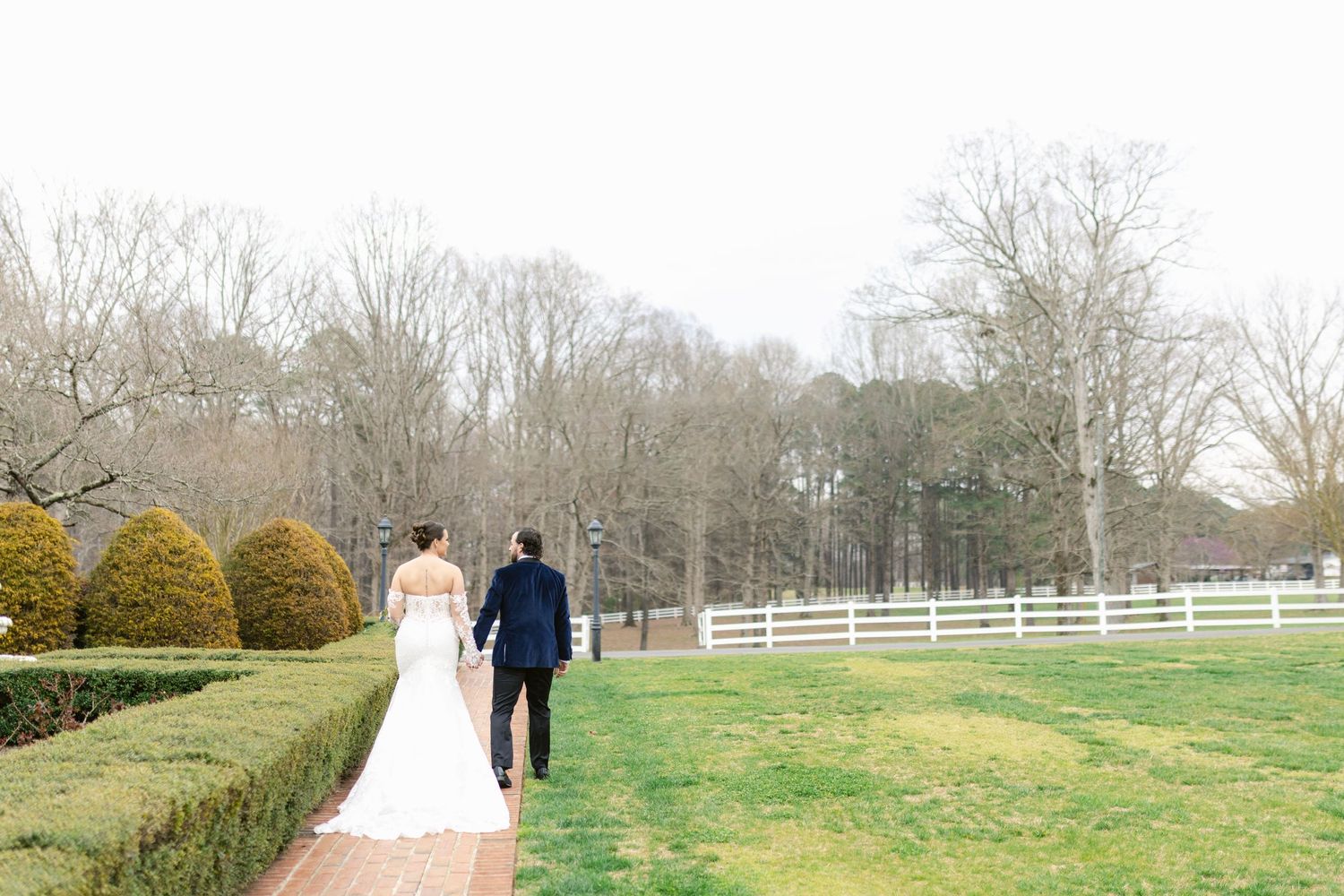 A couple walks along a brick path by white fenced grounds and manicured hedges at a scenic outdoor venue.