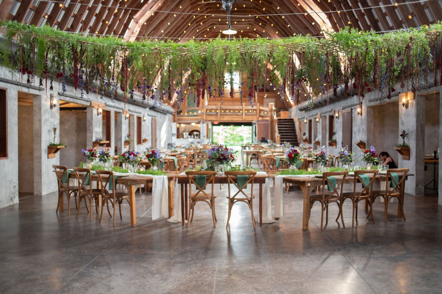 Rustic barn reception venue with vaulted ceiling, hanging greenery, and cross-back chairs.