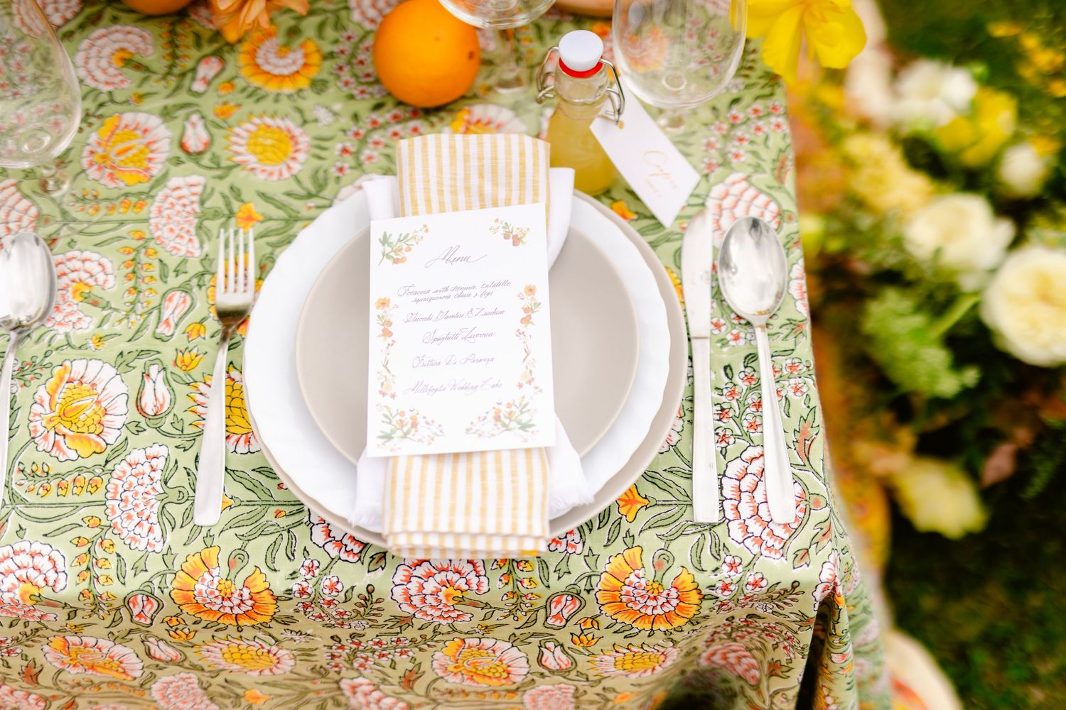Elegant table setting with citrus fruit and floral tablecloth featuring white plates and vintage-style silverware.