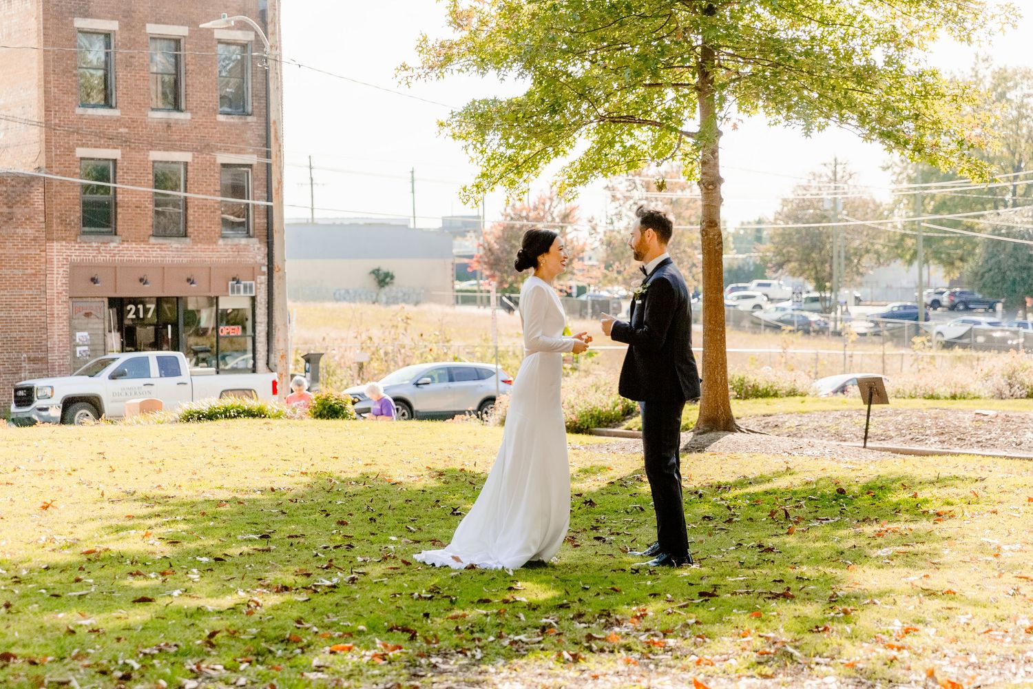 A couple in wedding attire stands on a grassy hill near brick buildings during golden hour.