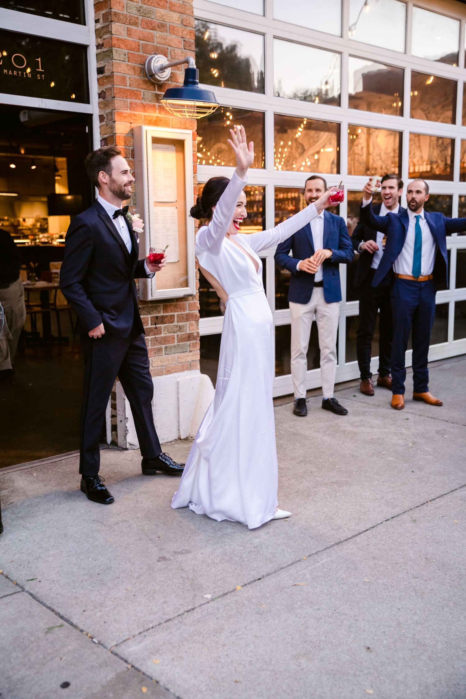 Wedding celebration outside a brick venue with guests cheering as the bride raises her arms in joy.