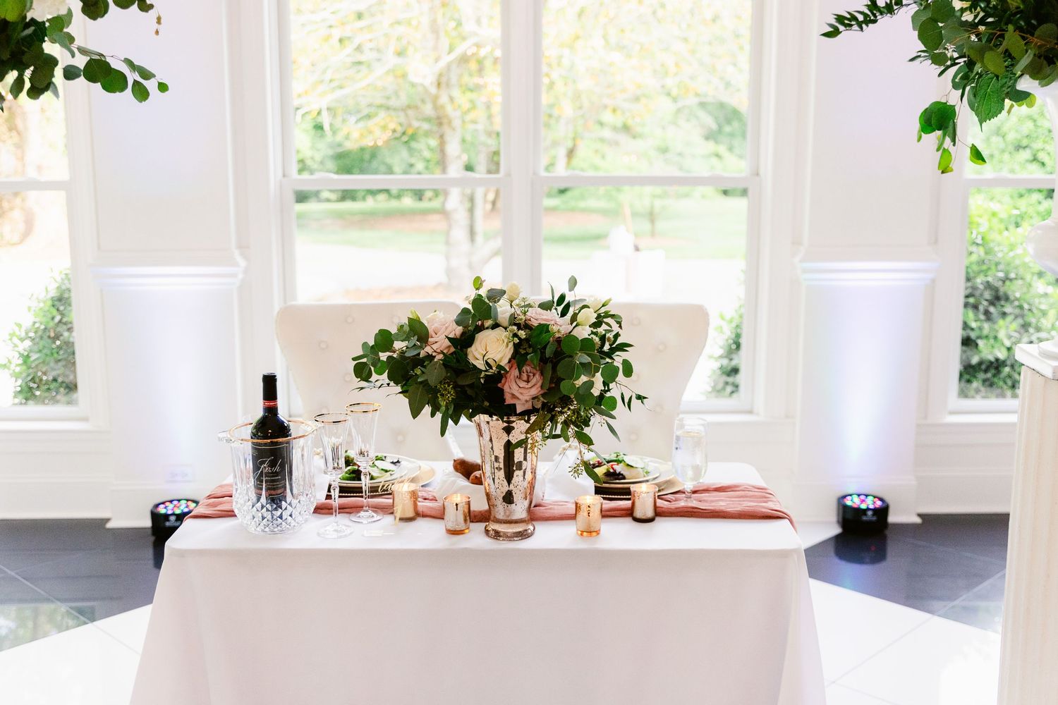 Elegant white dining room with large windows, natural light, and green foliage accents creating an airy atmosphere.