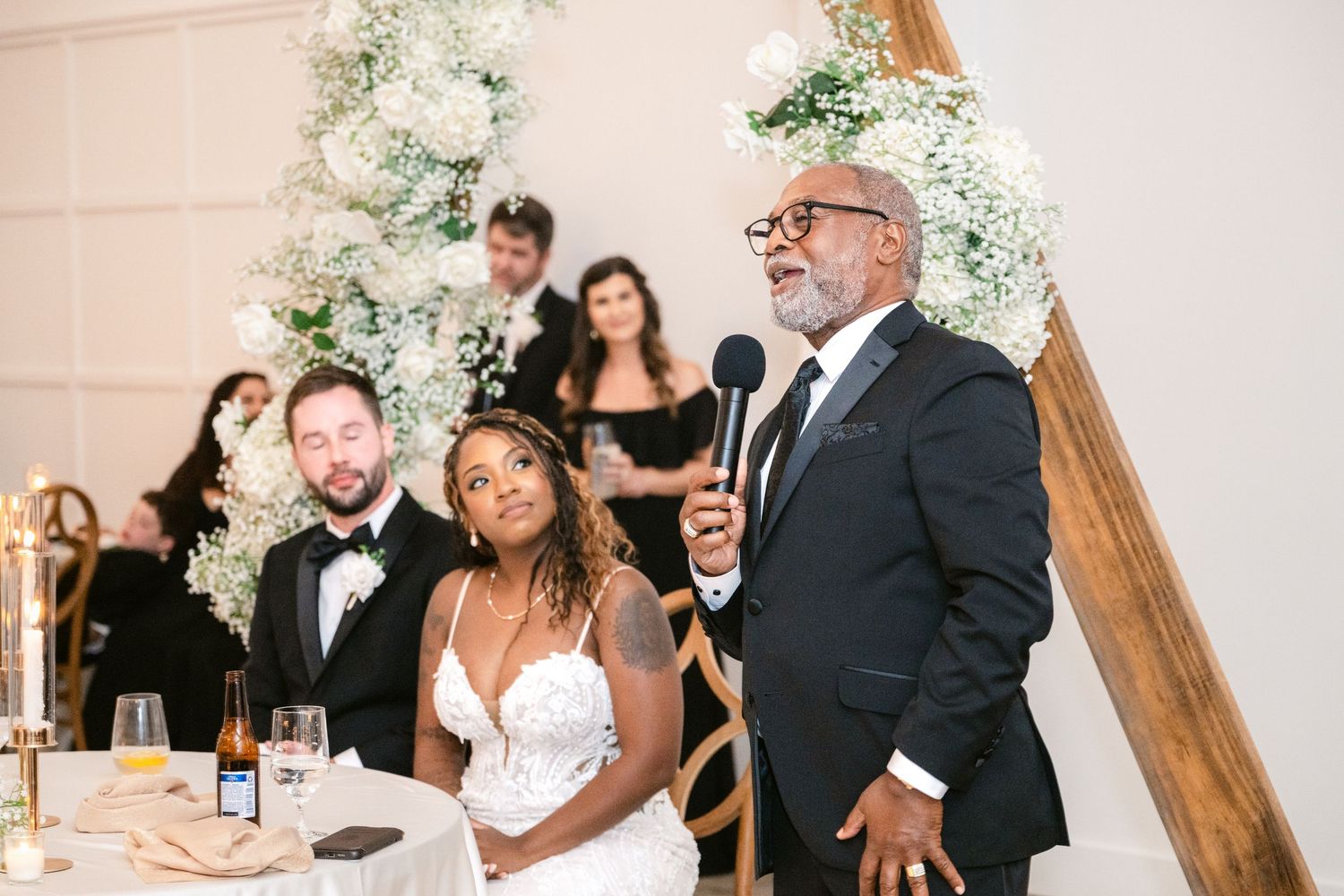 Wedding party celebrates with laughter and champagne during reception at elegant venue with white floral decor.