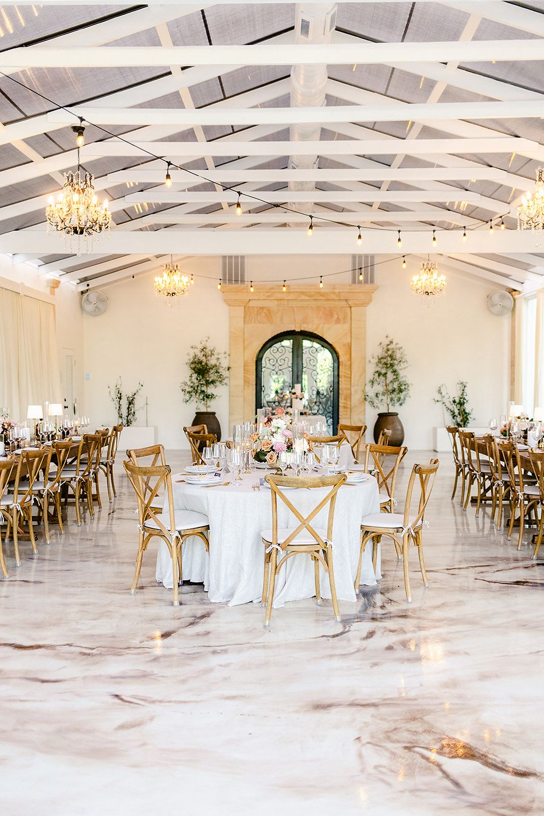 Elegant wedding reception venue with white vaulted ceiling, crystal chandeliers, and cross-back chairs at round tables.
