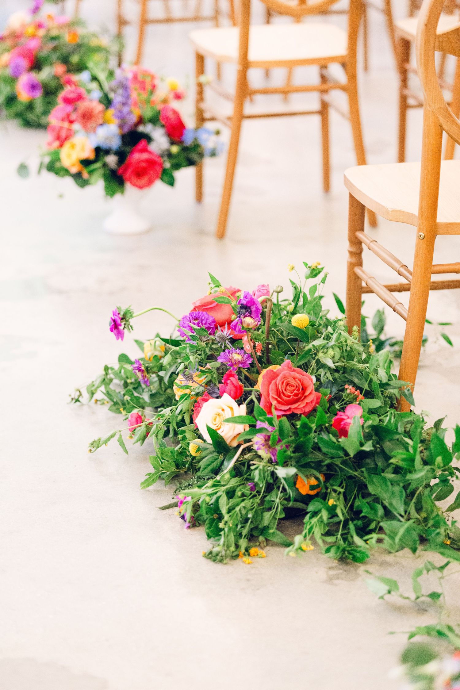 Vibrant pink and coral floral arrangements line a wedding aisle with lush greenery and wooden chairs.