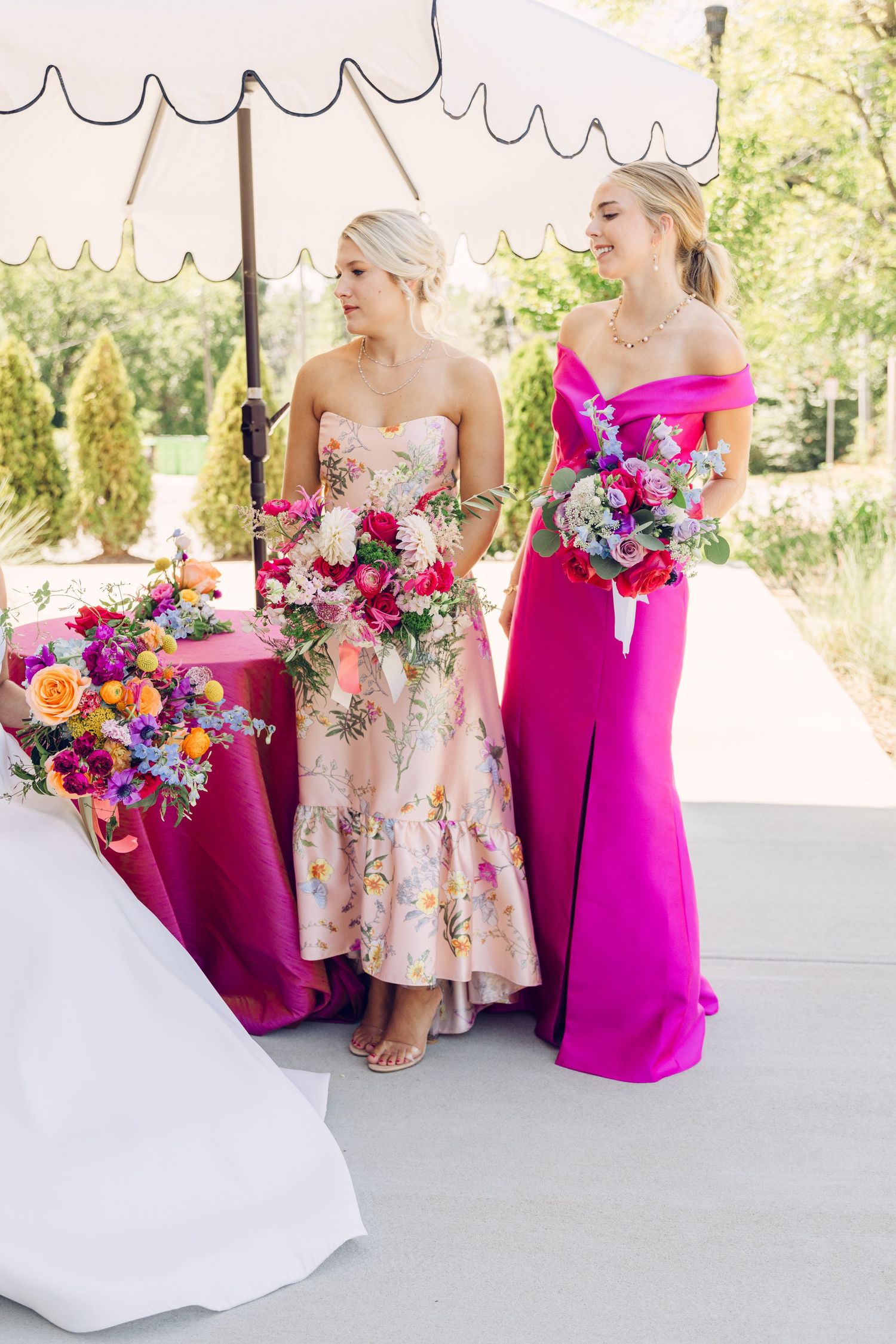 Bridesmaids in vibrant pink dresses hold colorful bouquets at an outdoor wedding venue.