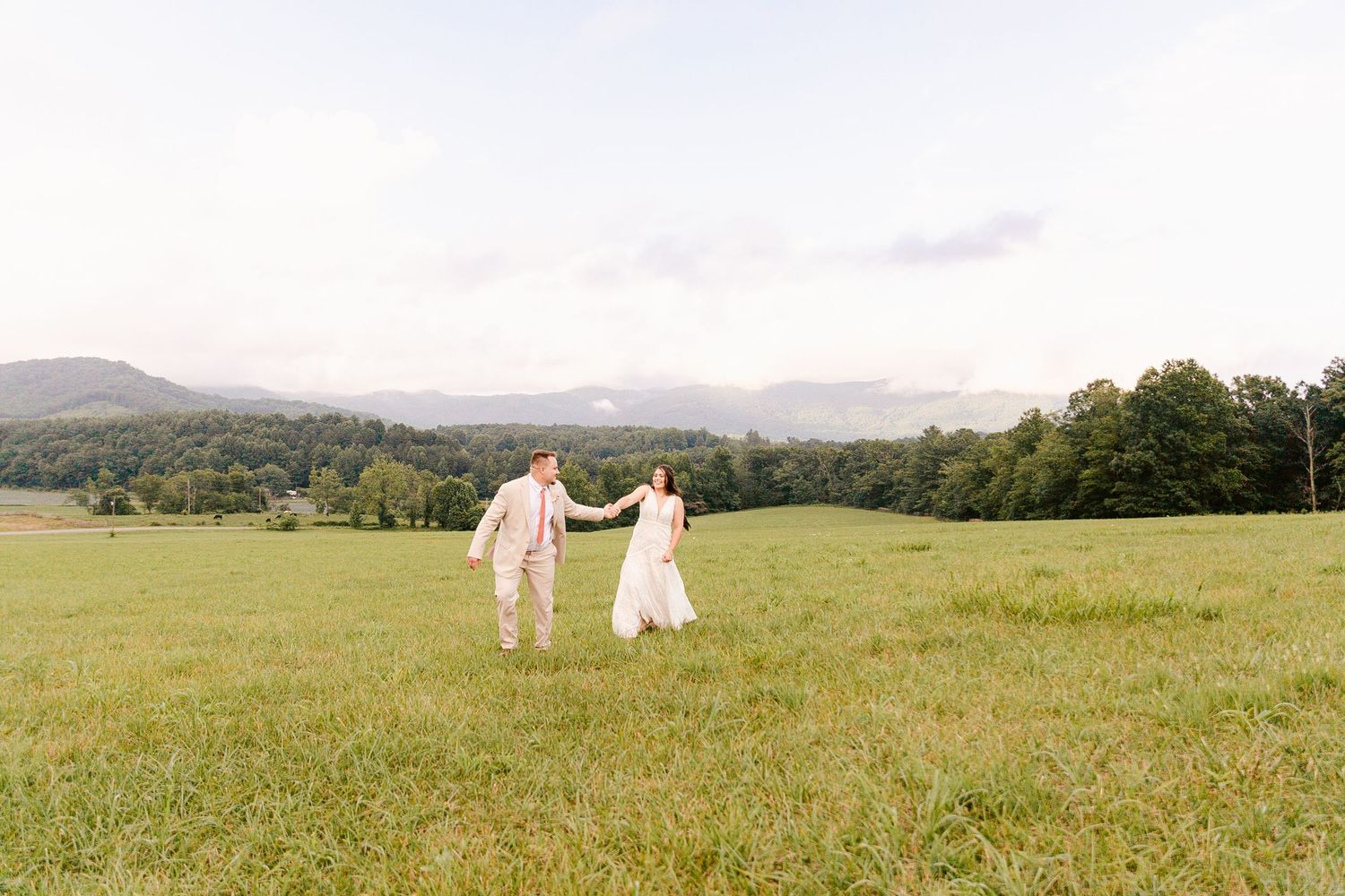 Two people in light-colored attire hold hands while walking through a scenic mountain meadow on a misty day.