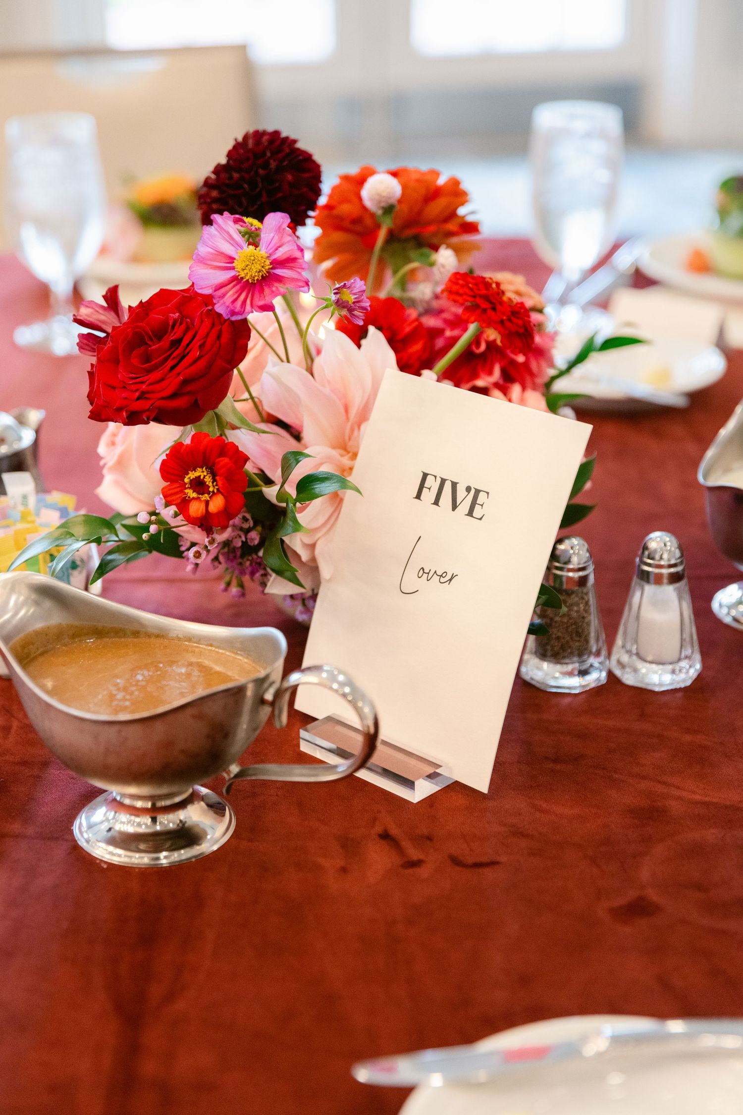 Red and pink floral centerpieces with table numbers and place settings on a polished wooden dining table.