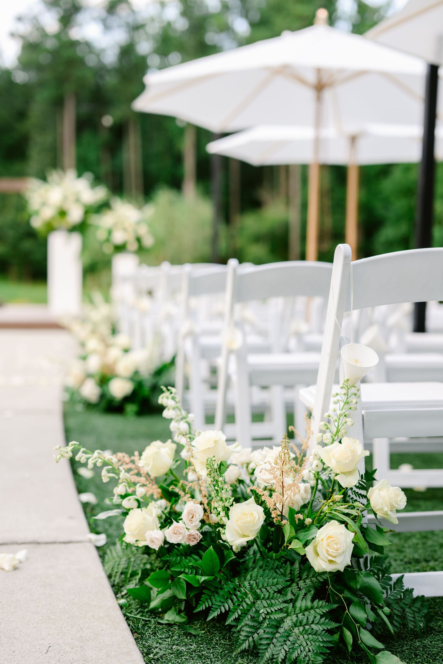 White garden chairs and elegant floral arrangements with roses and ferns line an outdoor wedding ceremony aisle.