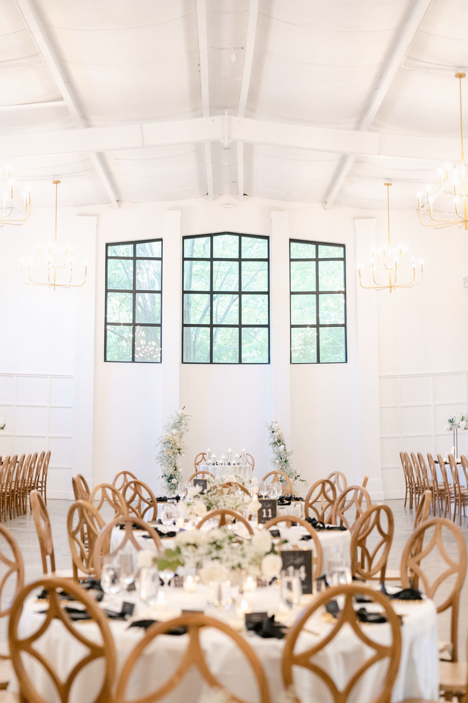 Modern white wedding reception venue with geometric wood chairs and elegant floral centerpieces under vaulted ceilings.