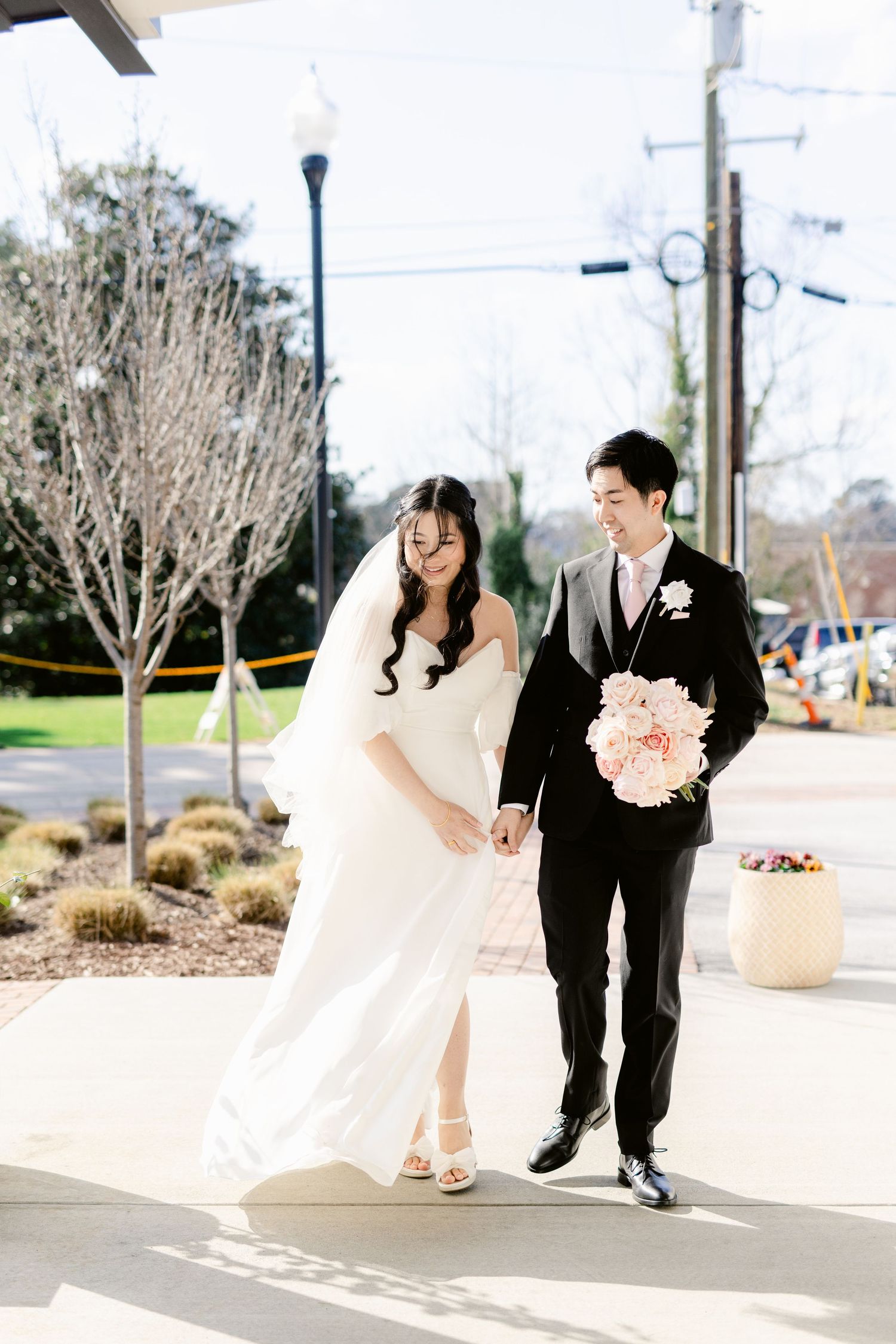 A couple in wedding attire walking and posing playfully on a street with pink bouquets.