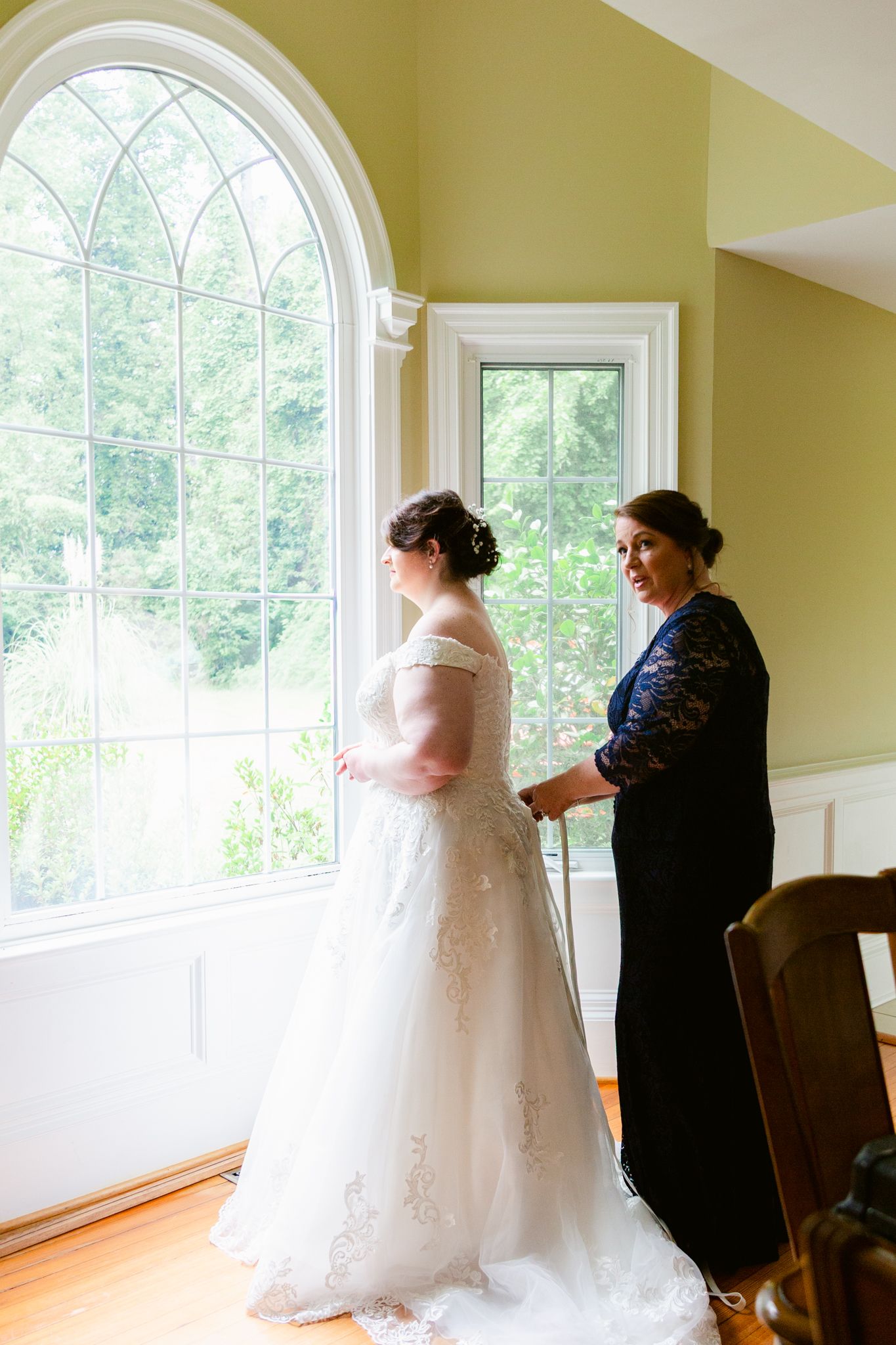 Two figures stand by an elegant arched window in a sunlit room while preparing for a formal occasion.