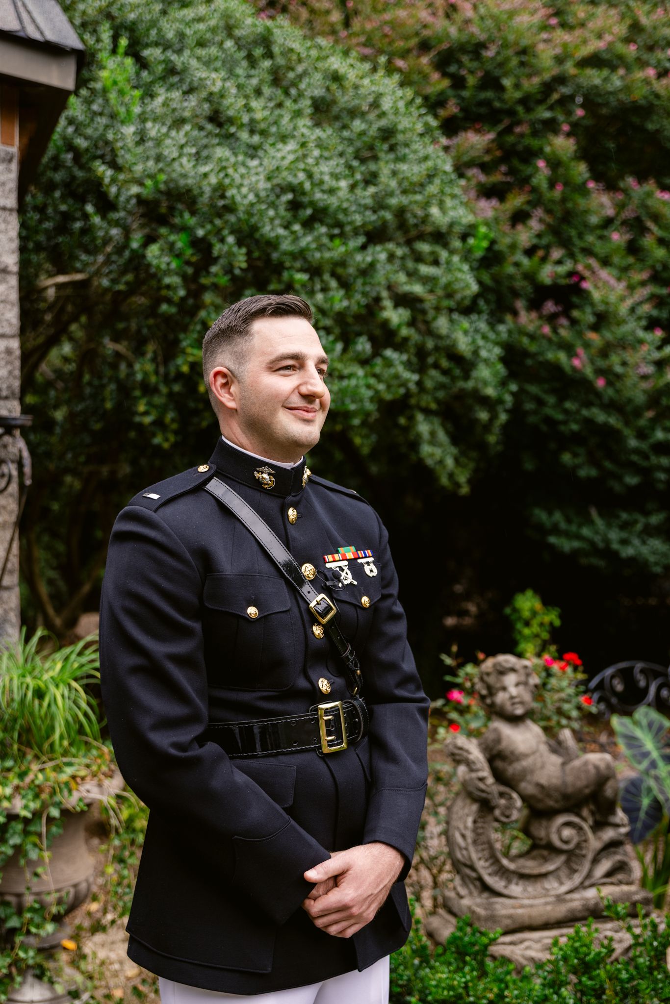 Military service member in dress uniform poses in a garden setting with decorative statues and greenery in the background.