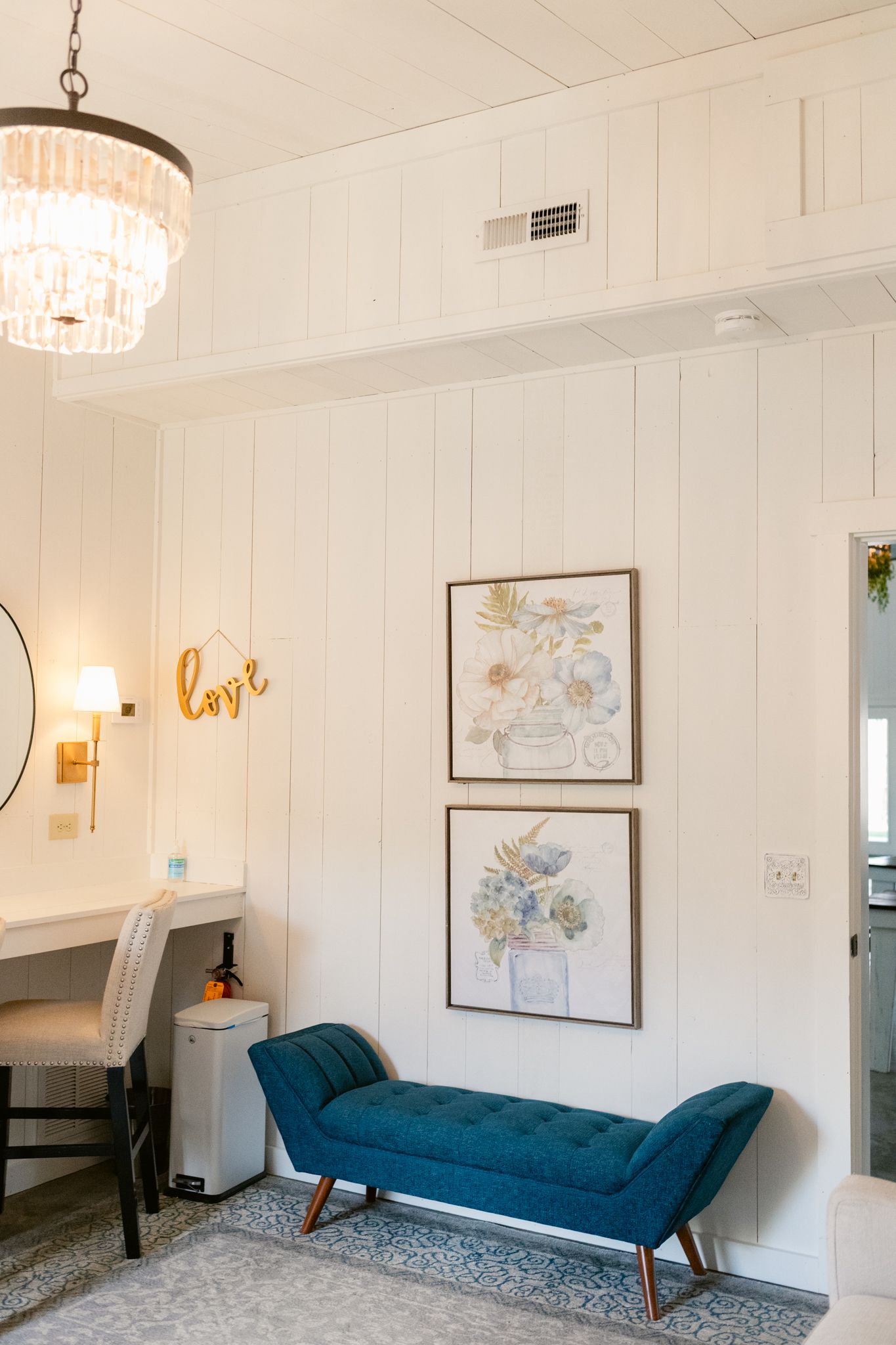 Modern white room interior with blue velvet chaise lounge, wall art, and crystal chandelier lighting.