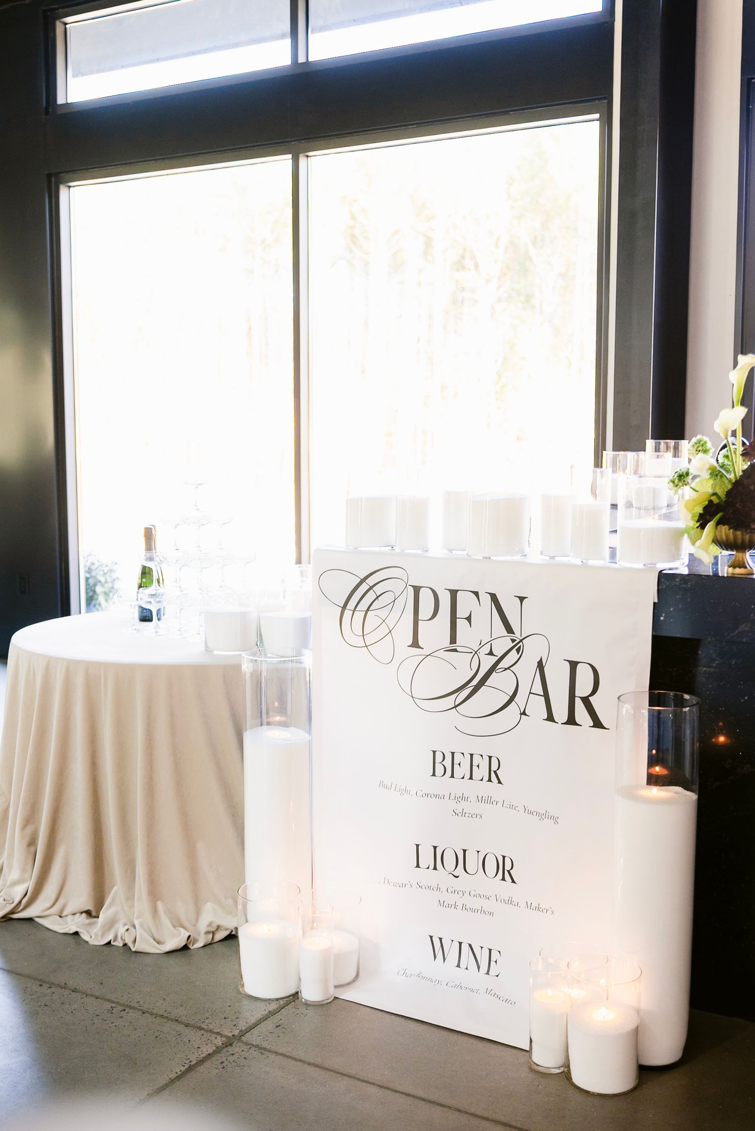 Elegant wedding bar setup with illuminated signs for wine, beer, and liquor displayed on white tablecloths with glowing candles.