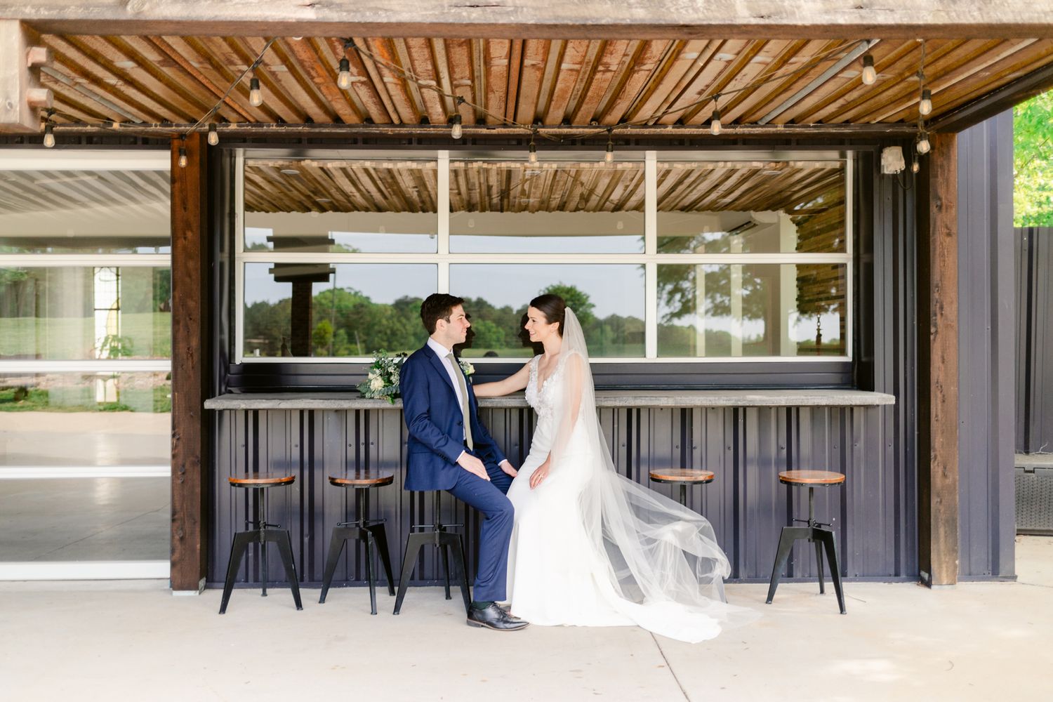 Couple in wedding attire shares a romantic moment under a modern covered walkway with wooden beams and glass panels.
