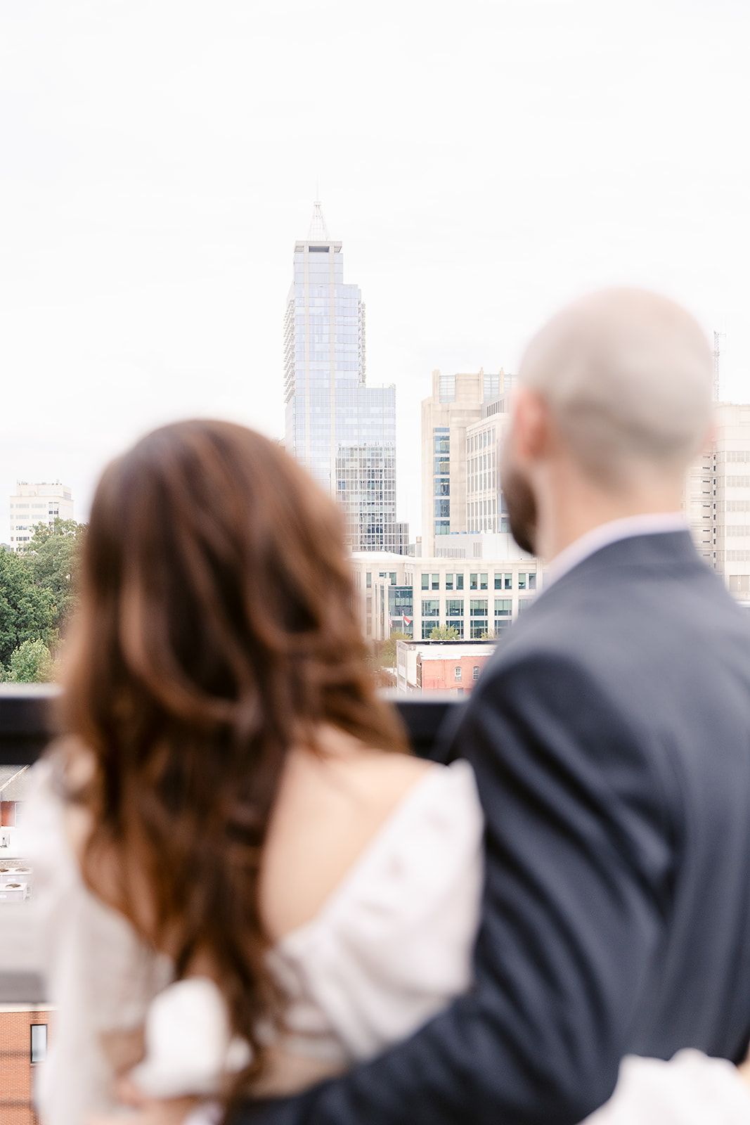 A couple in wedding attire stands together looking at a city skyline view from a rooftop.
