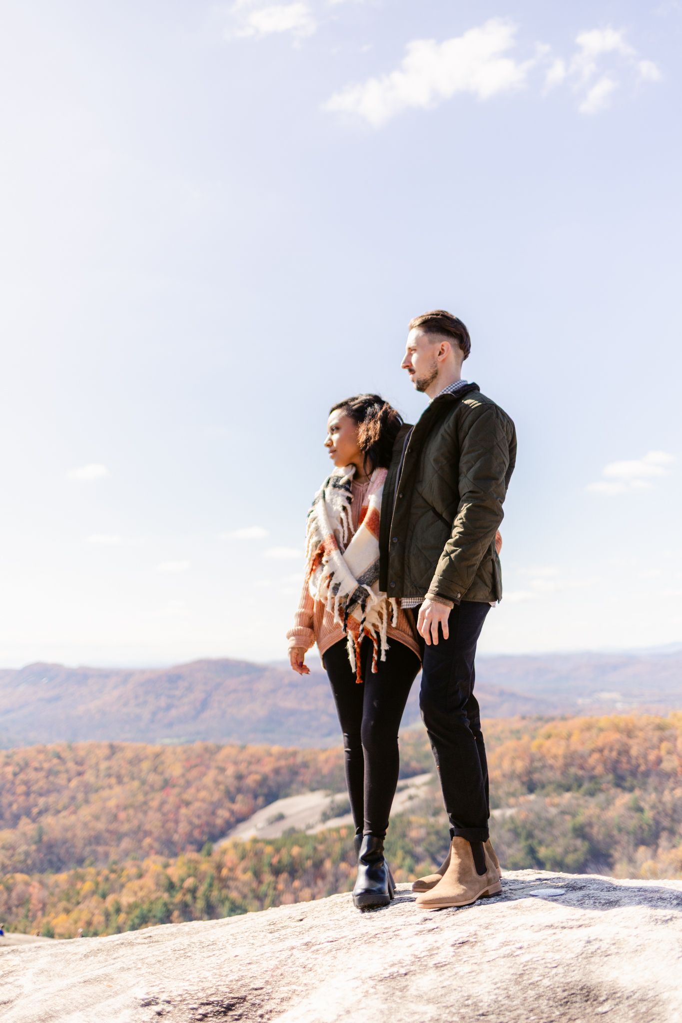 A couple stands together on a mountain overlook during autumn with colorful fall foliage in the valley below.