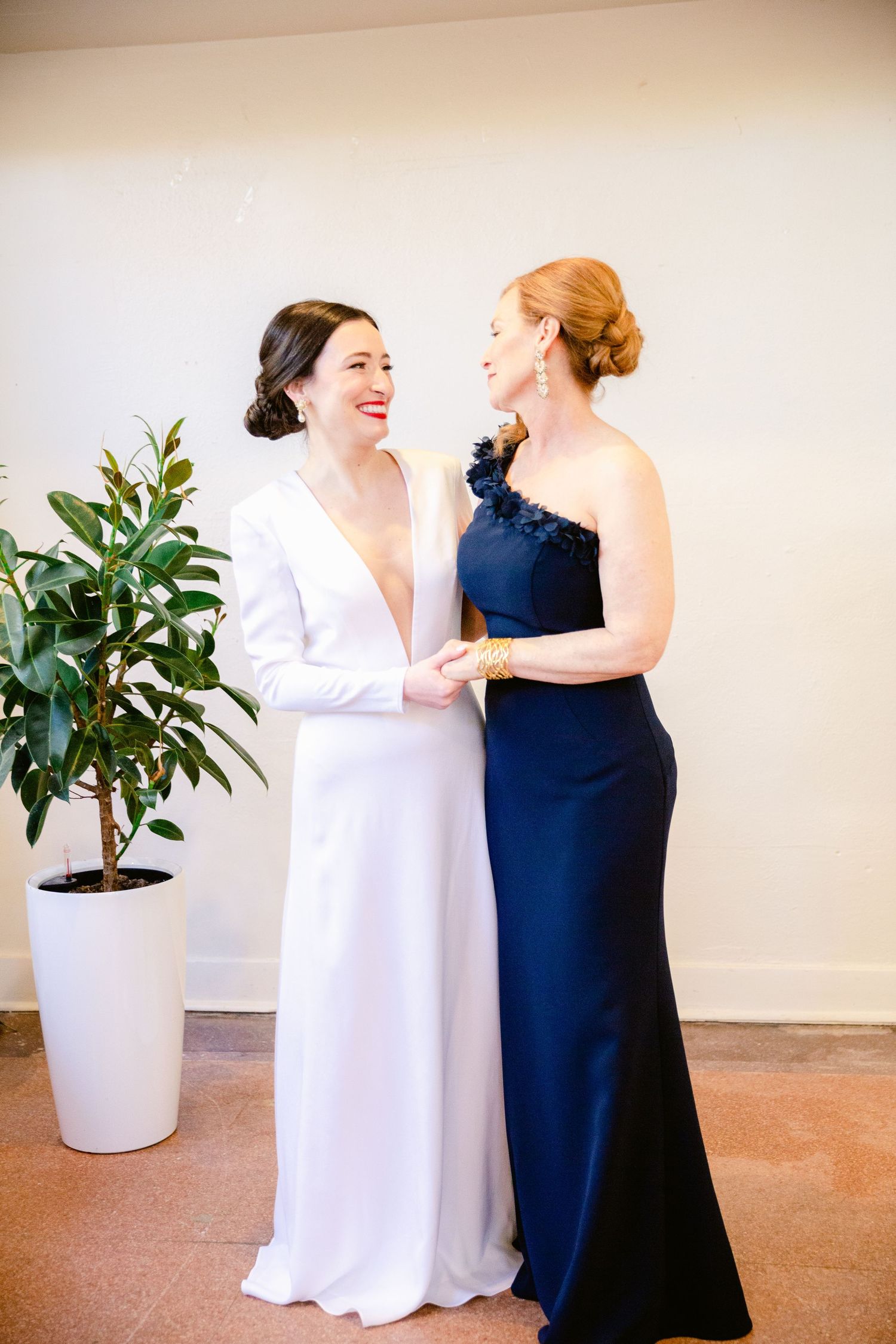 Two women in contrasting white and navy gowns share intimate moments in a minimalist setting with potted plants.
