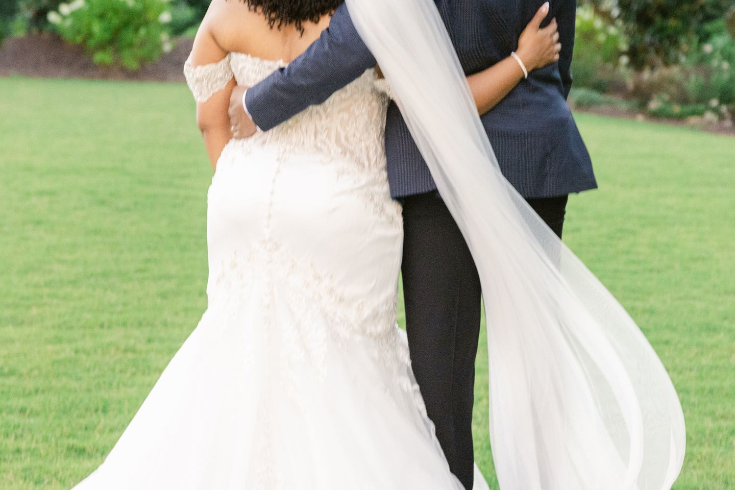 Newlywed couple embraces on a lush green lawn while the bride's veil flows elegantly in the breeze.
