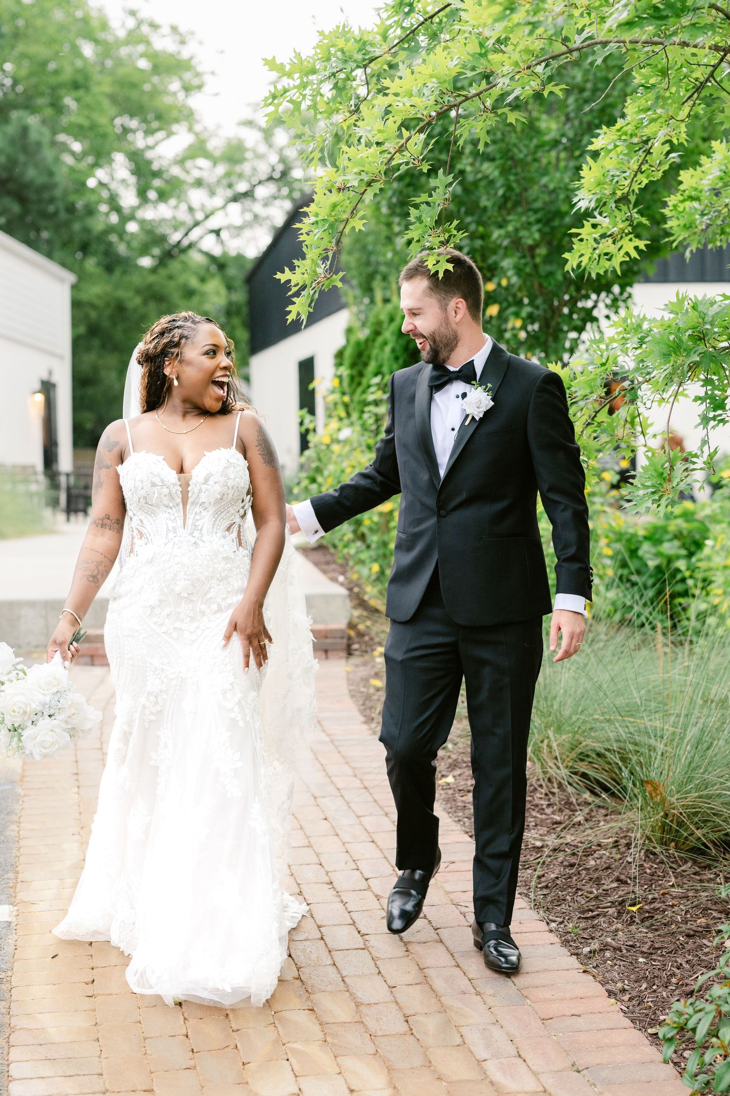 A couple walking and laughing together on a brick path surrounded by lush green gardens on their wedding day.